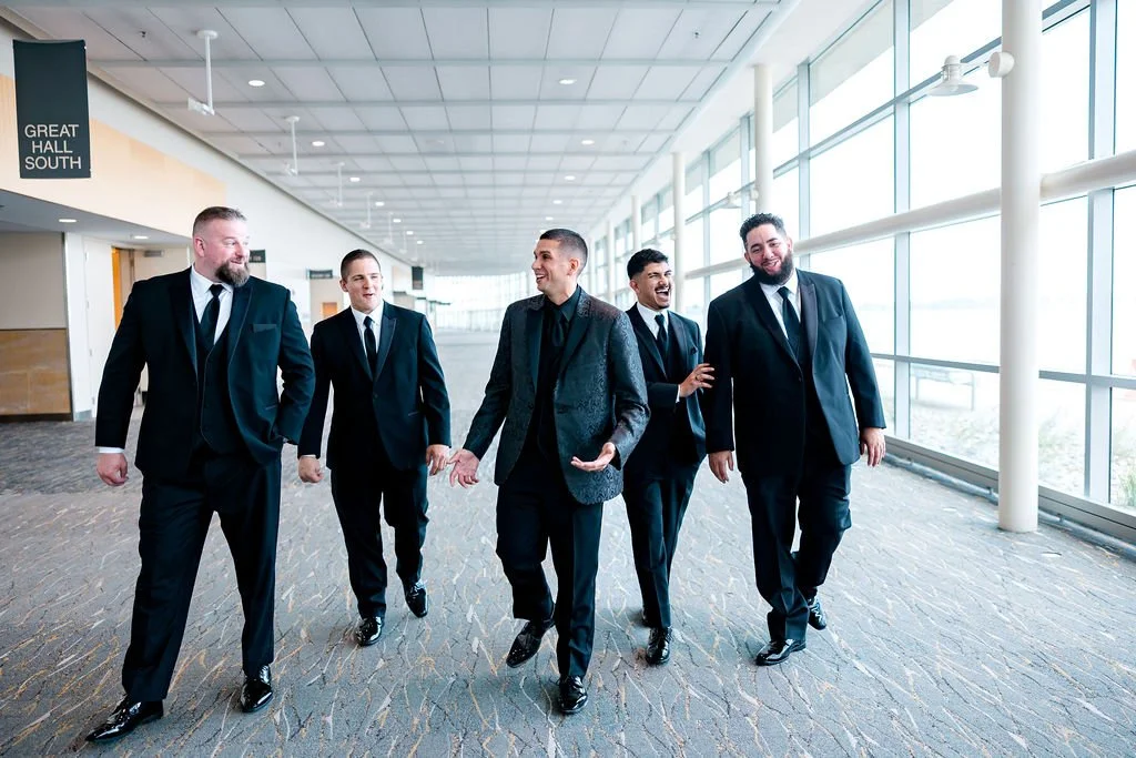 Group of five men in suits walking and chatting in a bright hallway with large windows.