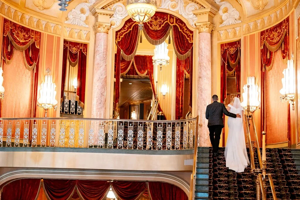 A couple holding hands at the top of a staircase inside a lavish, ornate theater with red curtains and golden decorations.