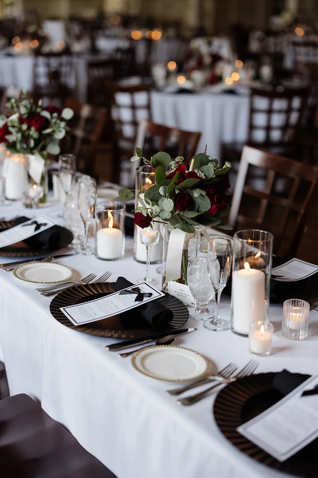Elegant banquet table decorated with candlelit centerpieces, red and white floral arrangements, black napkins, and gold-rimmed plates at an indoor event.