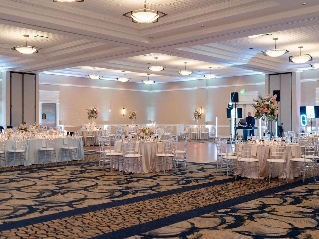 Event hall decorated for a wedding reception with round tables, white chairs, floral centerpieces, and a DJ setup.