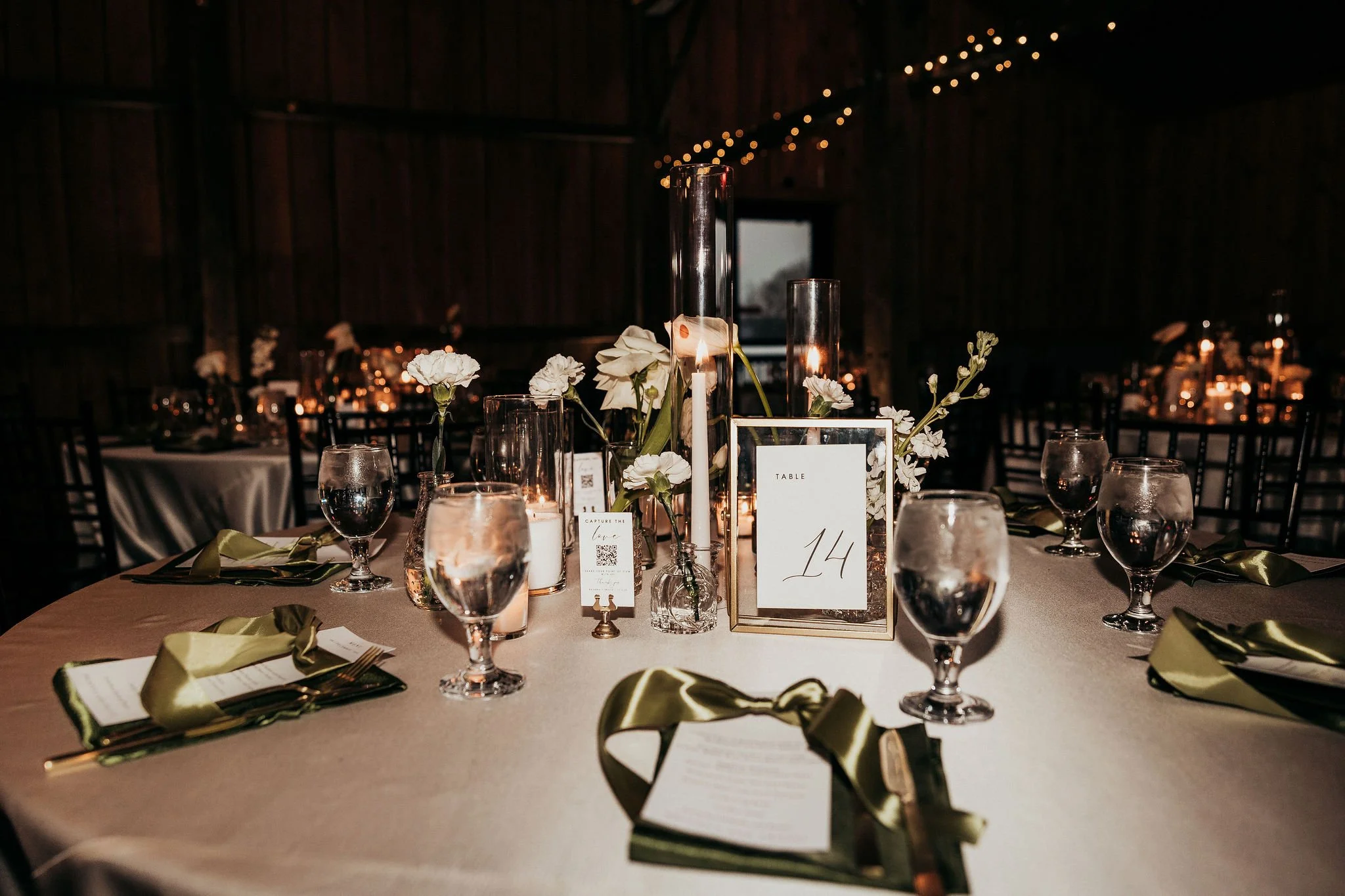 Wedding reception table decorated with white flowers, candles in glass holders, and table number 14, set in a rustic barn with string lights overhead.