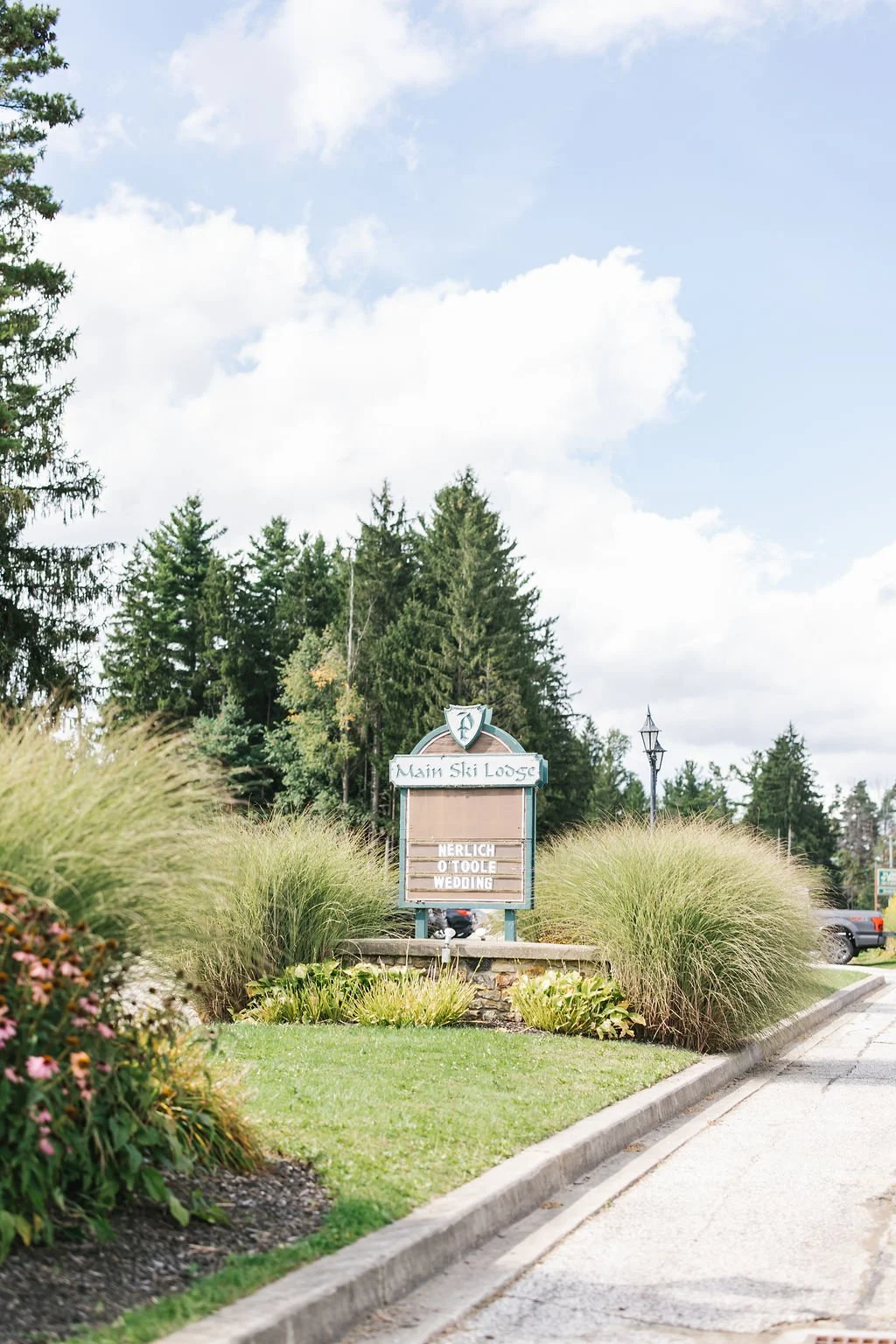 A sign for Main Ski Lodge surrounded by tall grasses and greenery, with a forest of tall trees and a partly cloudy sky in the background.