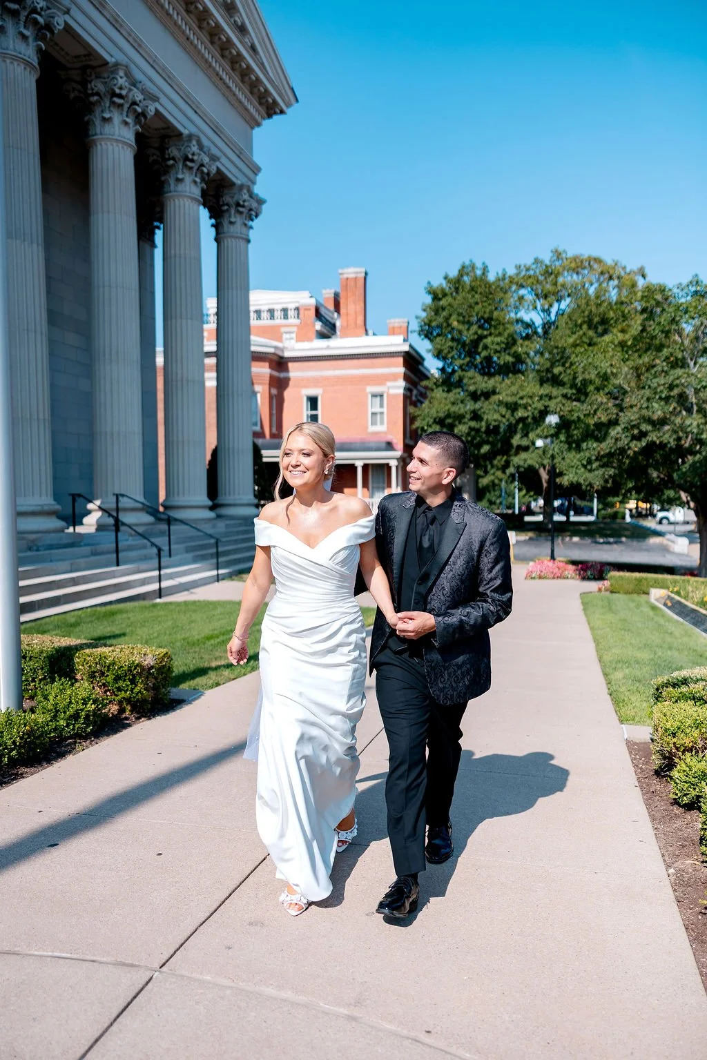 A bride and groom walking arm-in-arm on a sidewalk outside a historic building with large columns, on a sunny day with clear blue skies.