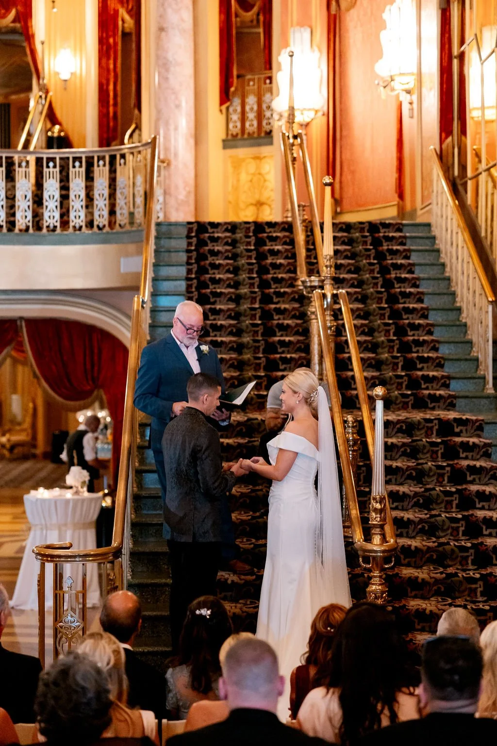 A couple getting married on a grand staircase with a officiant reading from a book, surrounded by guests seated in a lavish, elegant venue with ornate drapes and decor.