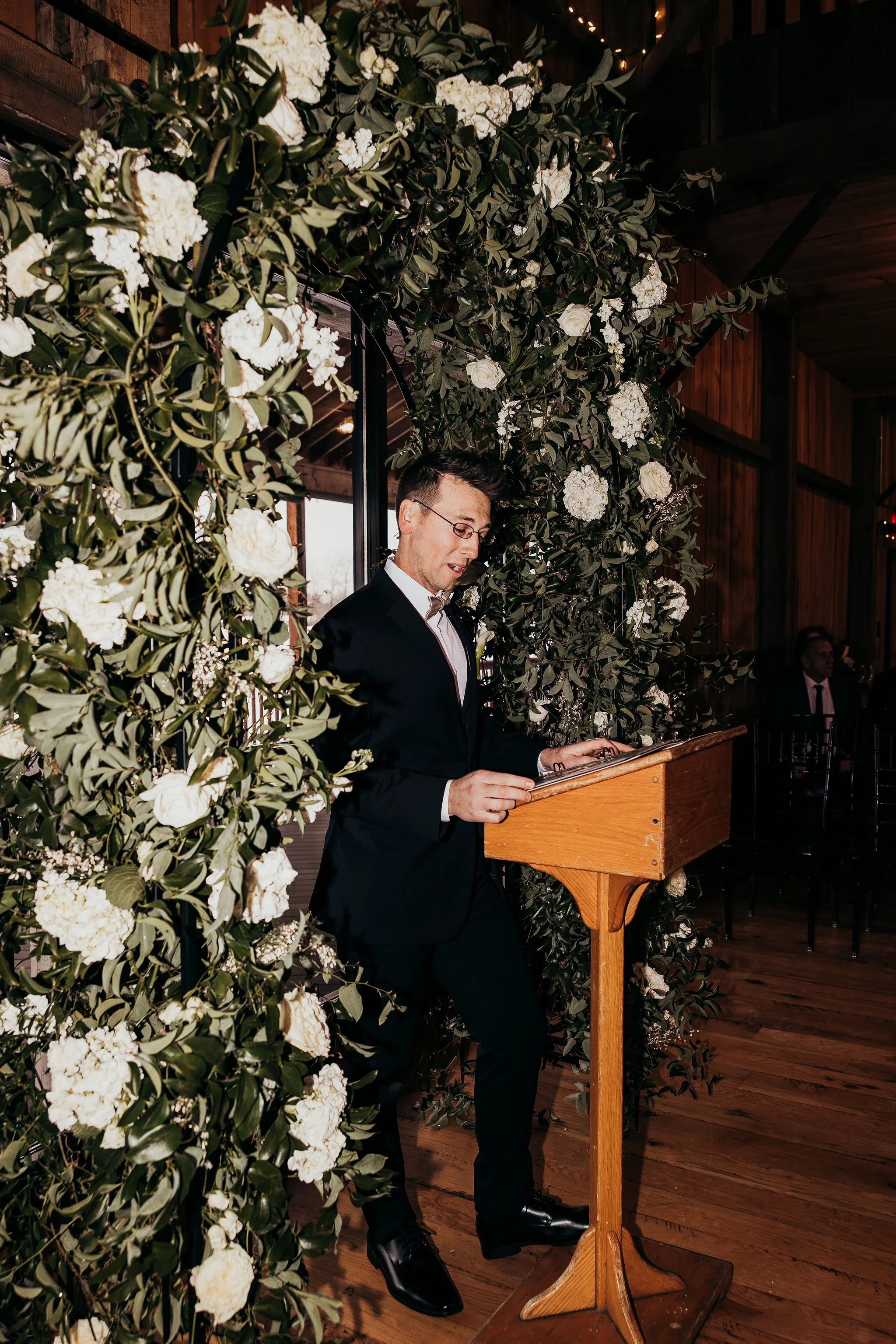 A man in a black tuxedo and glasses is standing at a wooden podium reading or speaking in front of a large floral arrangement of white flowers and green leaves at a formal event indoors with wooden walls and ceiling.