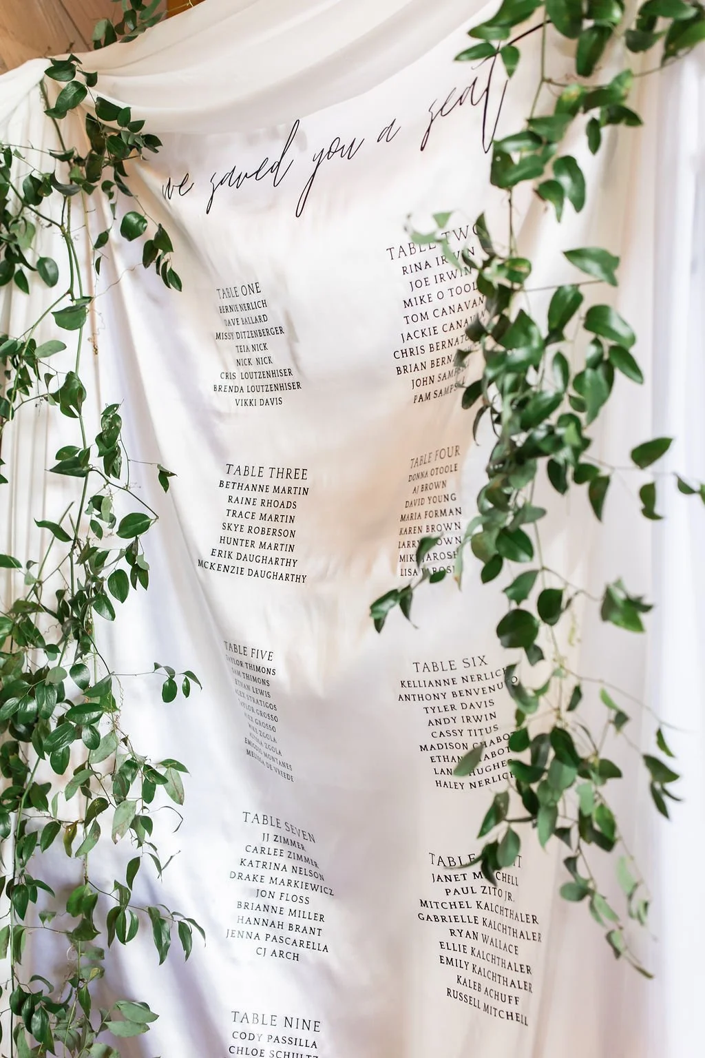 Wedding seating chart with handwritten calligraphy on a white fabric backdrop decorated with green ivy.