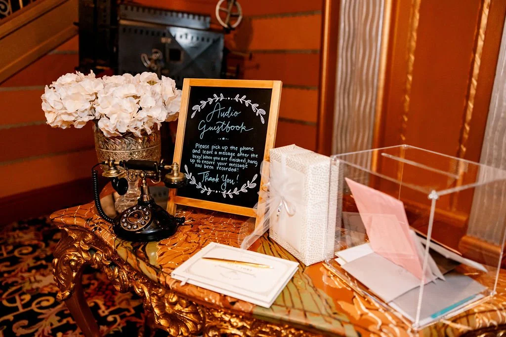 Vintage black rotary telephone, floral arrangement with white flowers, framed blackboard sign, gift box, clear donation box, and stationery on ornate wooden table.