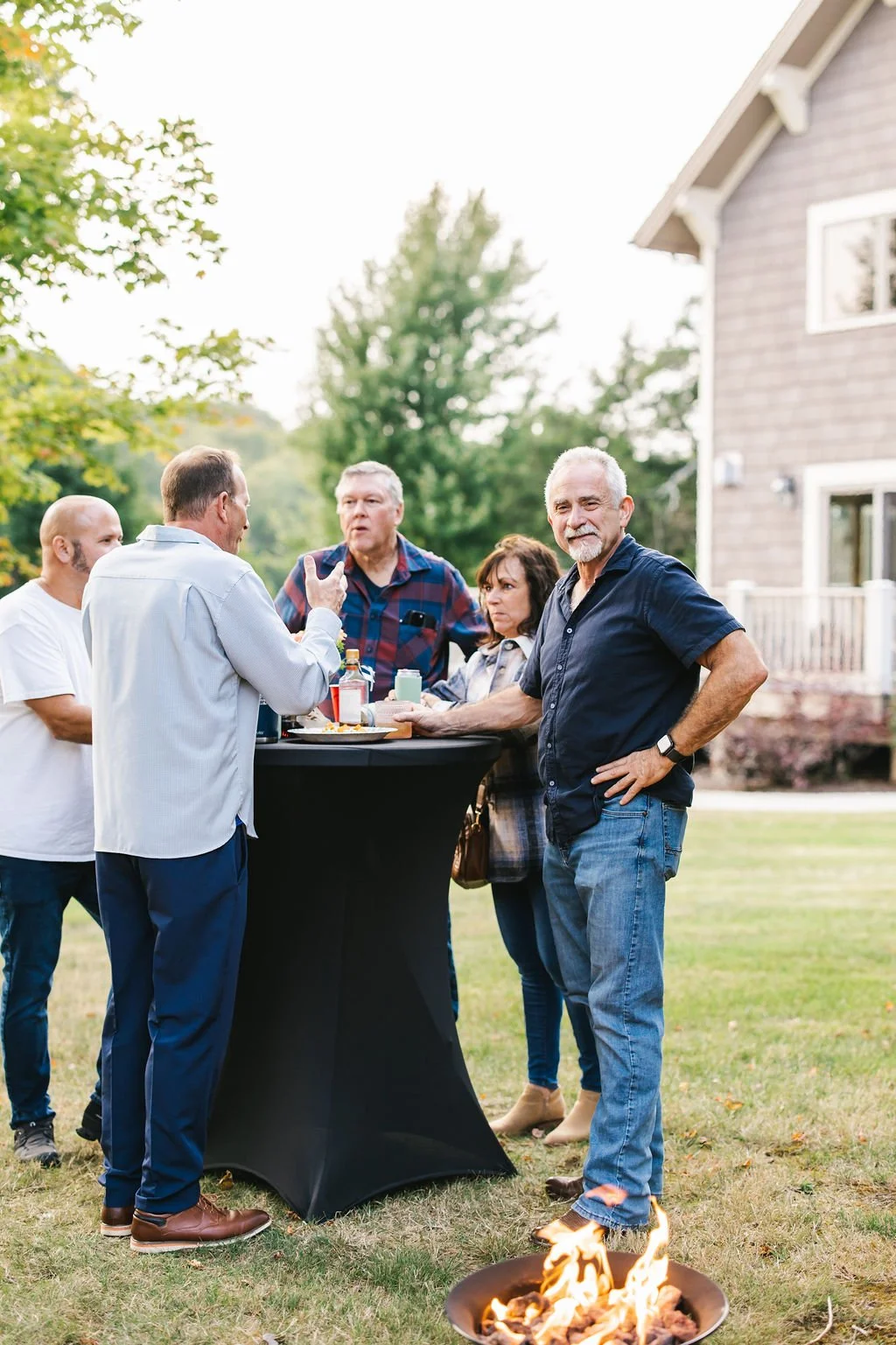 A group of five adults socializing and enjoying drinks outdoors near a tall black table on a lawn, with a small fire pit nearby, in front of a house with trees and a blue sky in the background.