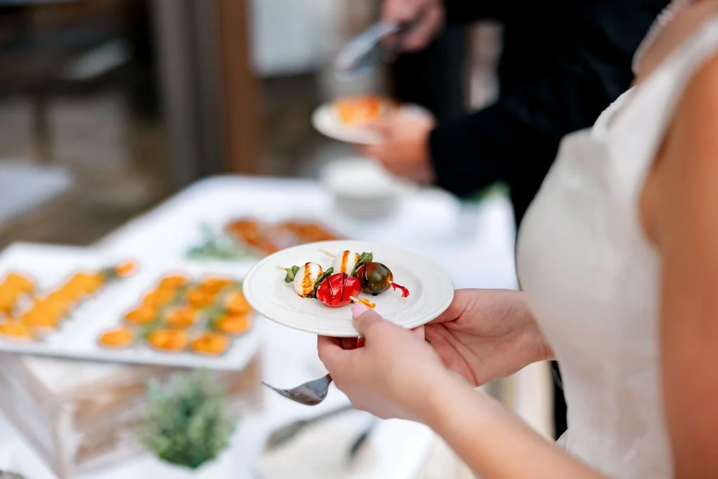 Person holding a small white plate with colorful appetizers, with a buffet table of similar dishes in the background.