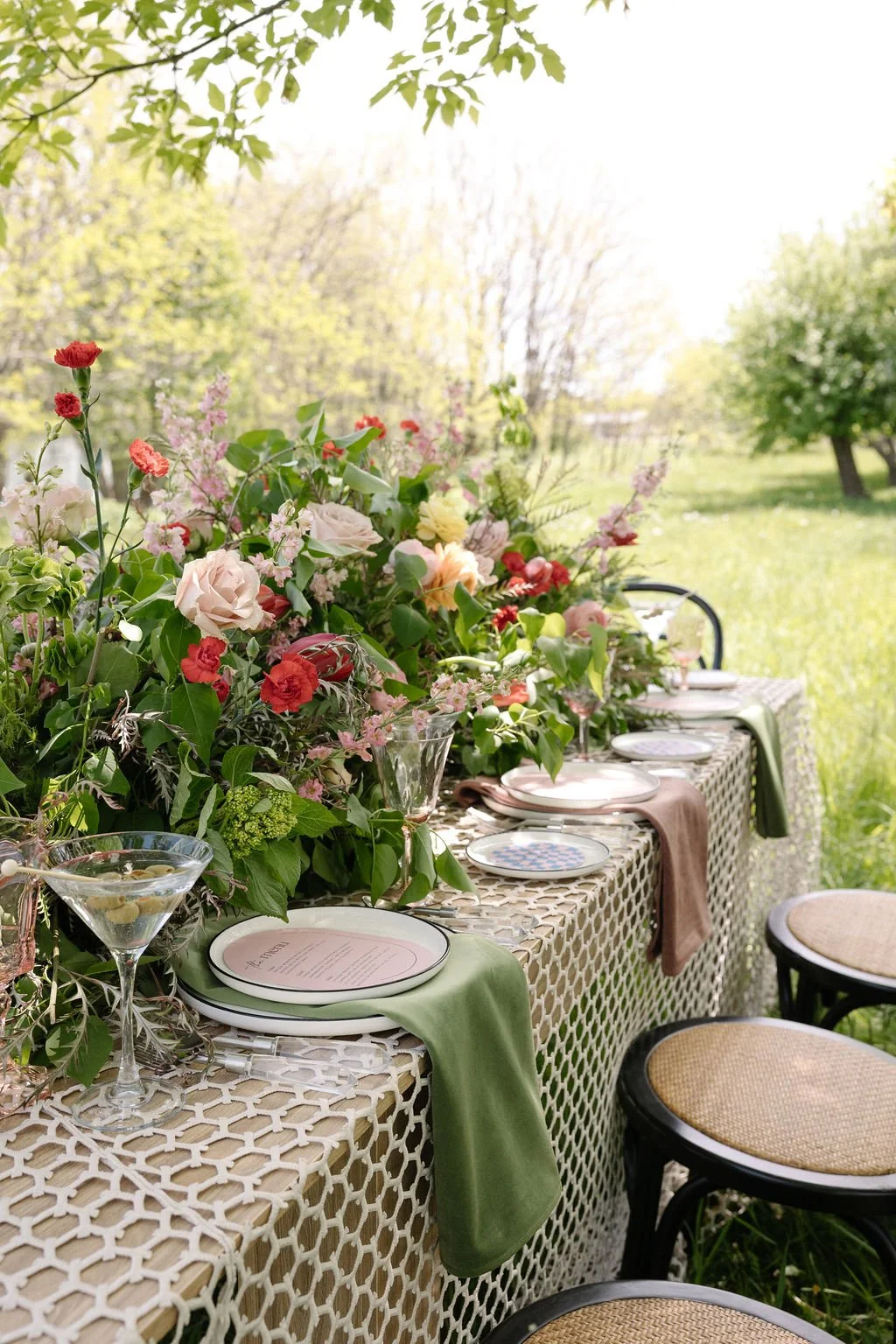 Outdoor dining table decorated with flowers, plates, glasses, and napkins, set in a garden with trees and green grass