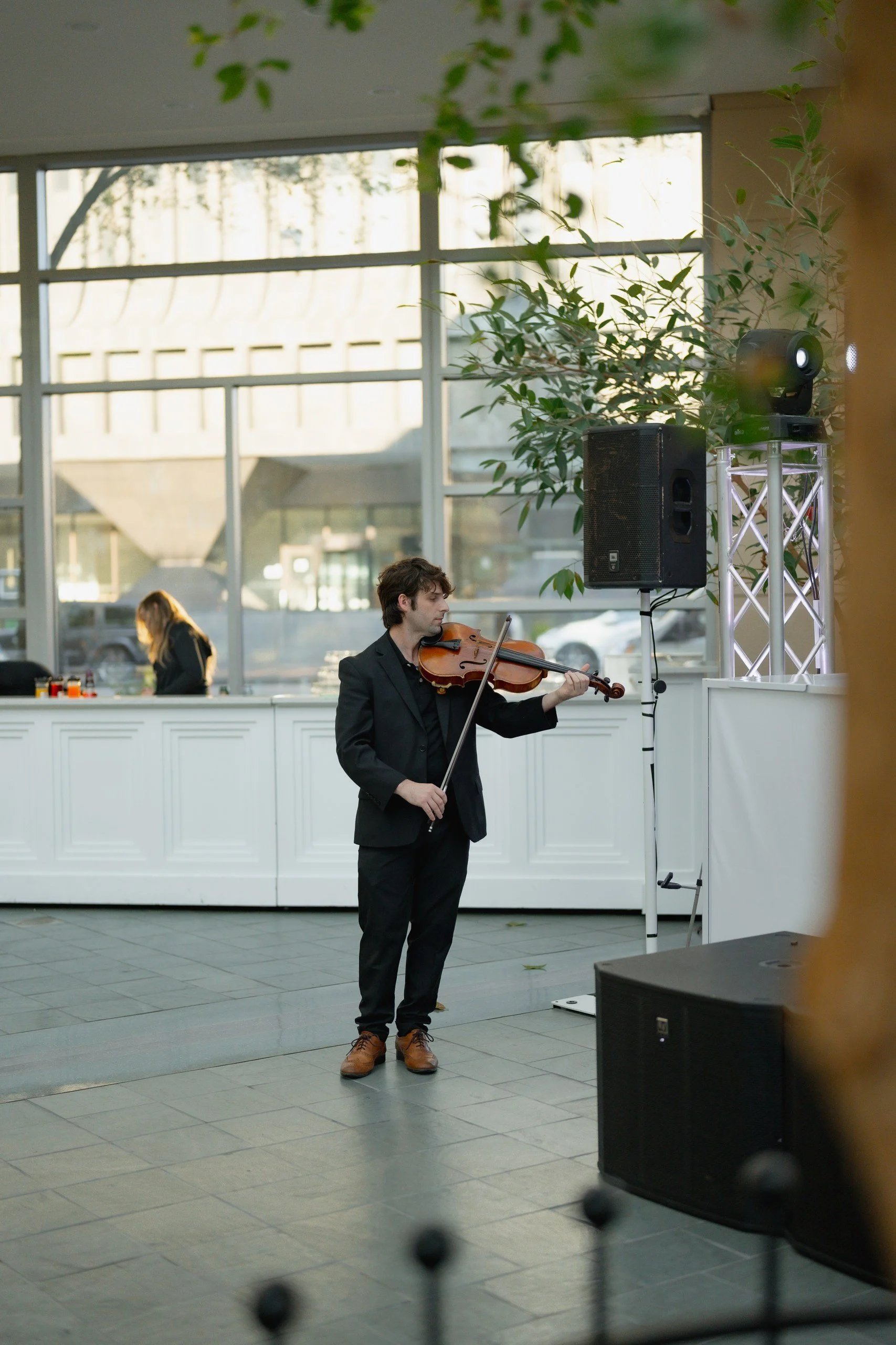 A man in a black suit playing a violin indoors during a performance, with two large black speakers and a lighting equipment stand nearby, and a woman in the background behind a white counter, inside a building with large window panes and trees outsid