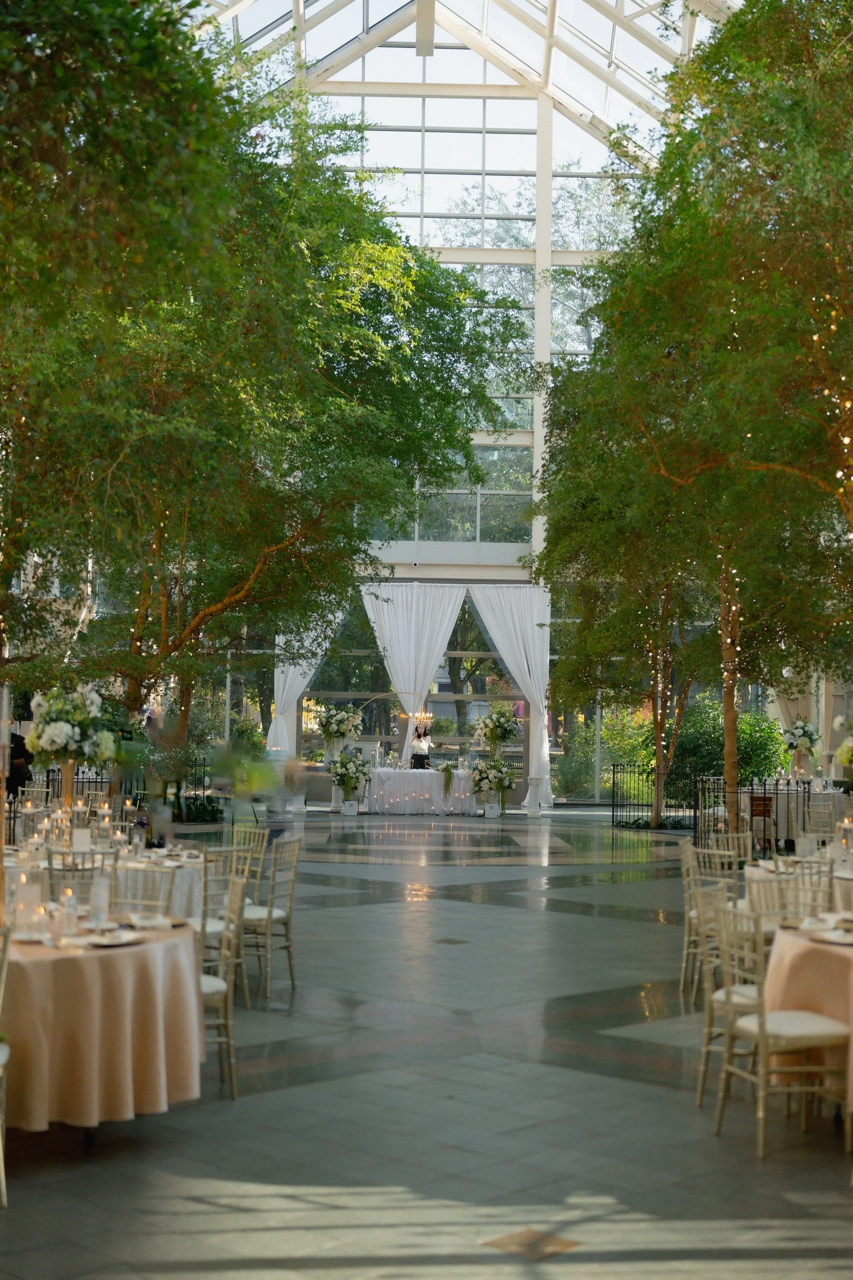 Indoor wedding reception setup inside a glass-walled venue with tall trees, white drapery, floral arrangements, and round tables with chairs.