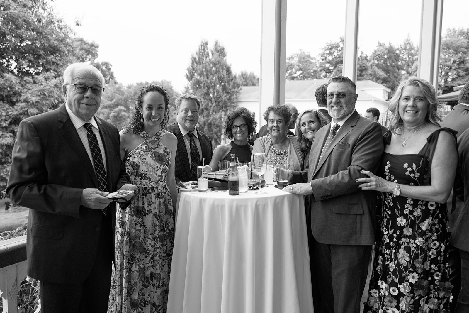 Black and white photo of eight people dressed in formal attire standing around a high cocktail table with drinks, smiling at the camera. The setting appears to be indoors with large windows and an outdoor landscape visible in the background.