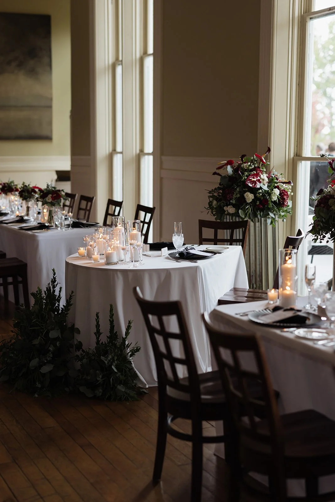 Elegant dining setup with tables covered in white tablecloths, decorated with candles, glassware, and floral arrangements in a well-lit room with large windows.