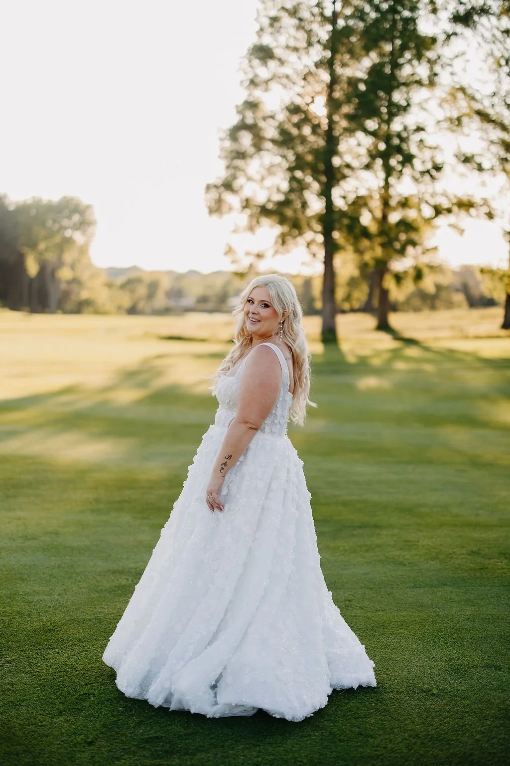 A woman in a white wedding dress standing on a grassy field with trees in the background, during sunset.