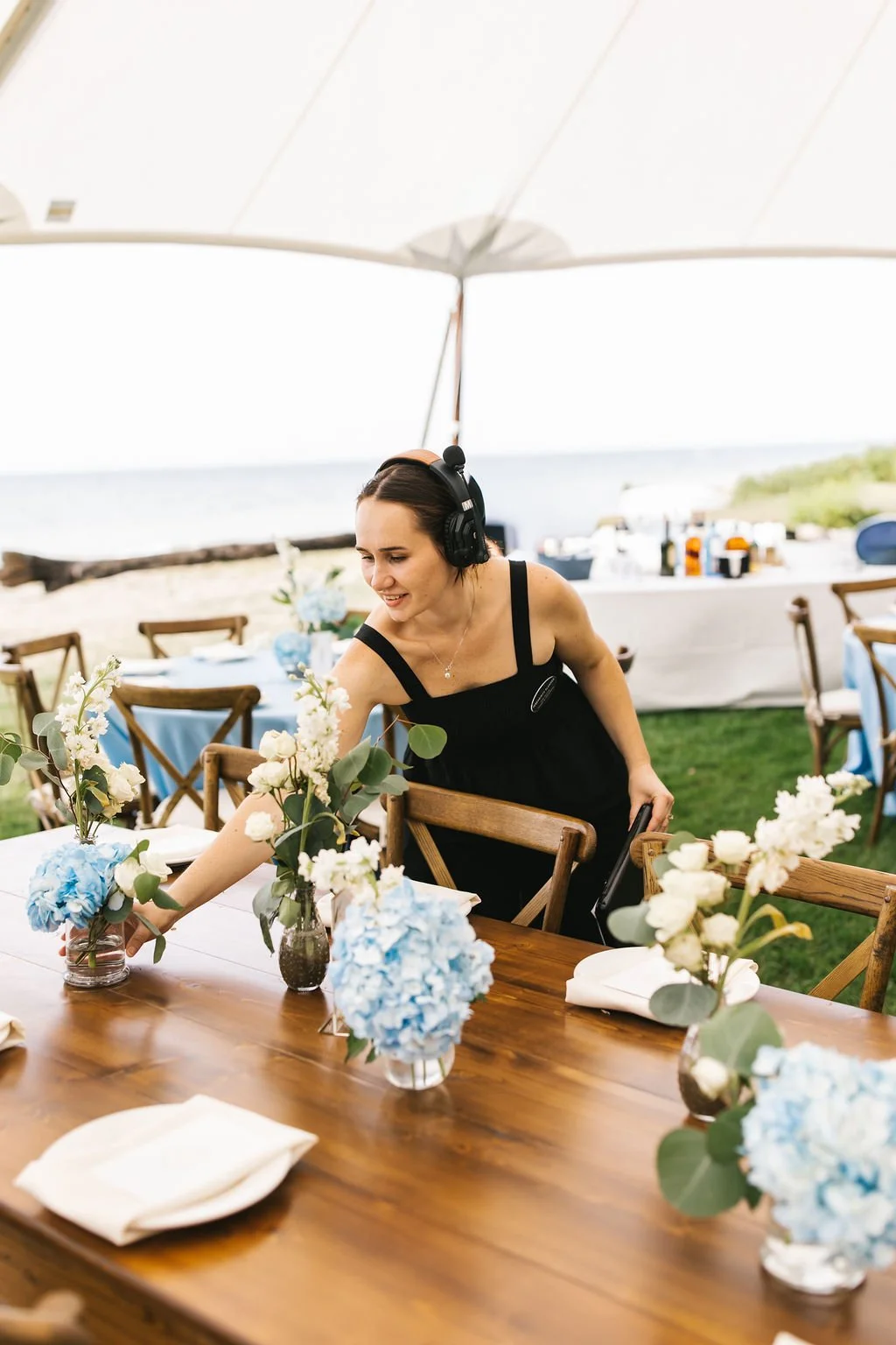 A woman wearing a black dress and headphones arranging flowers on a dining table outdoors near the beach.