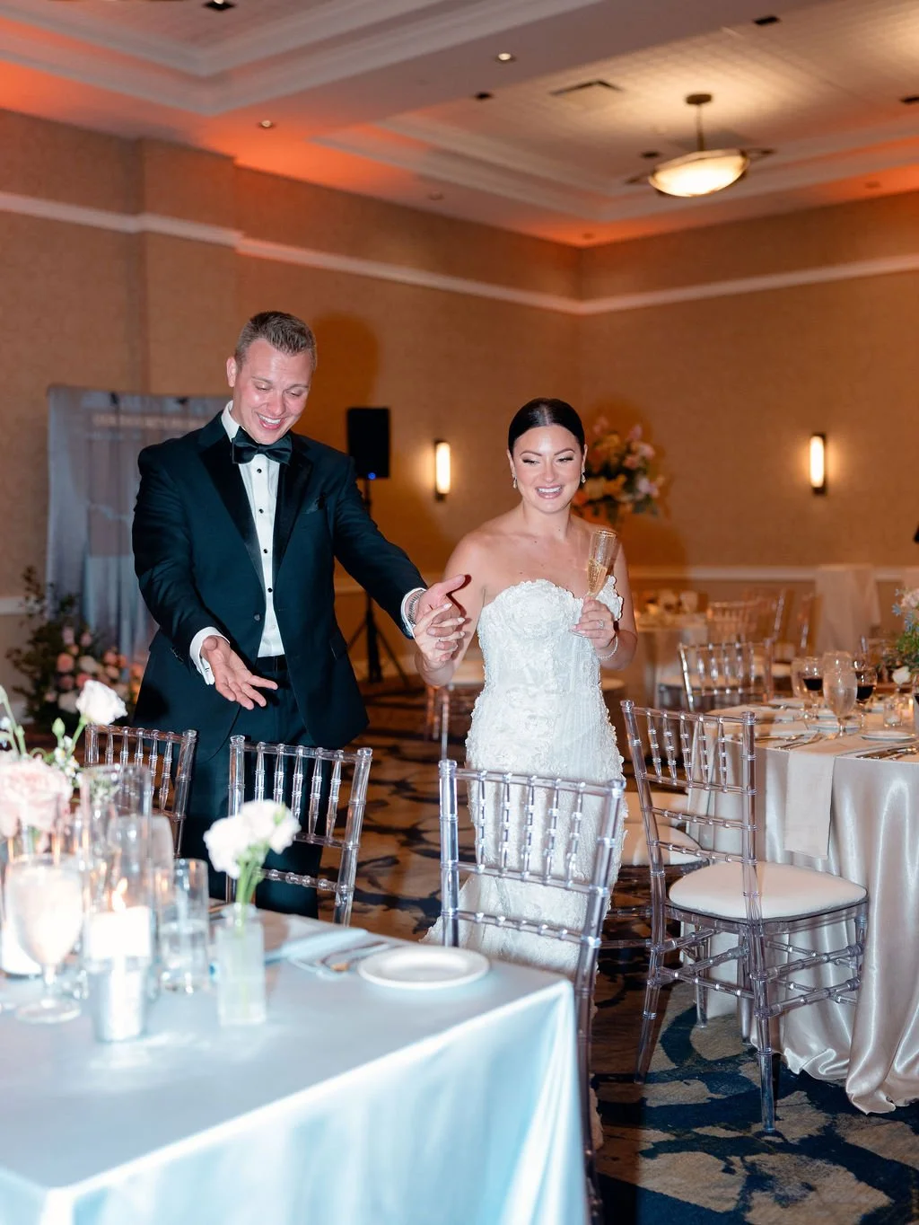 A newlywed couple in formal wedding attire walking and smiling through a decorated banquet hall, with the bride holding a champagne flute.