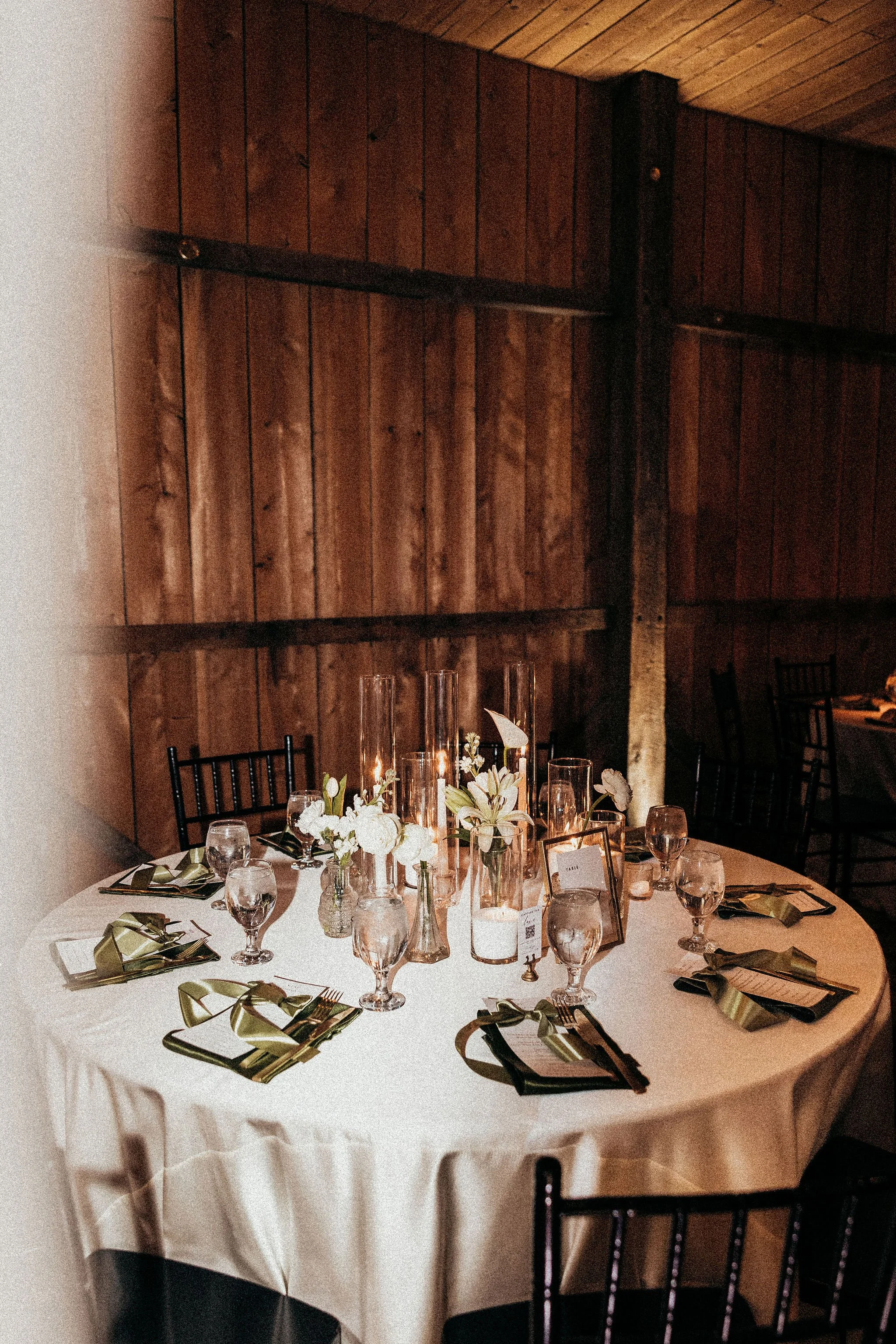A round table set for a formal event with a white tablecloth, floral centerpiece, and various glasses, plates, and utensils, inside a rustic wood-paneled room.