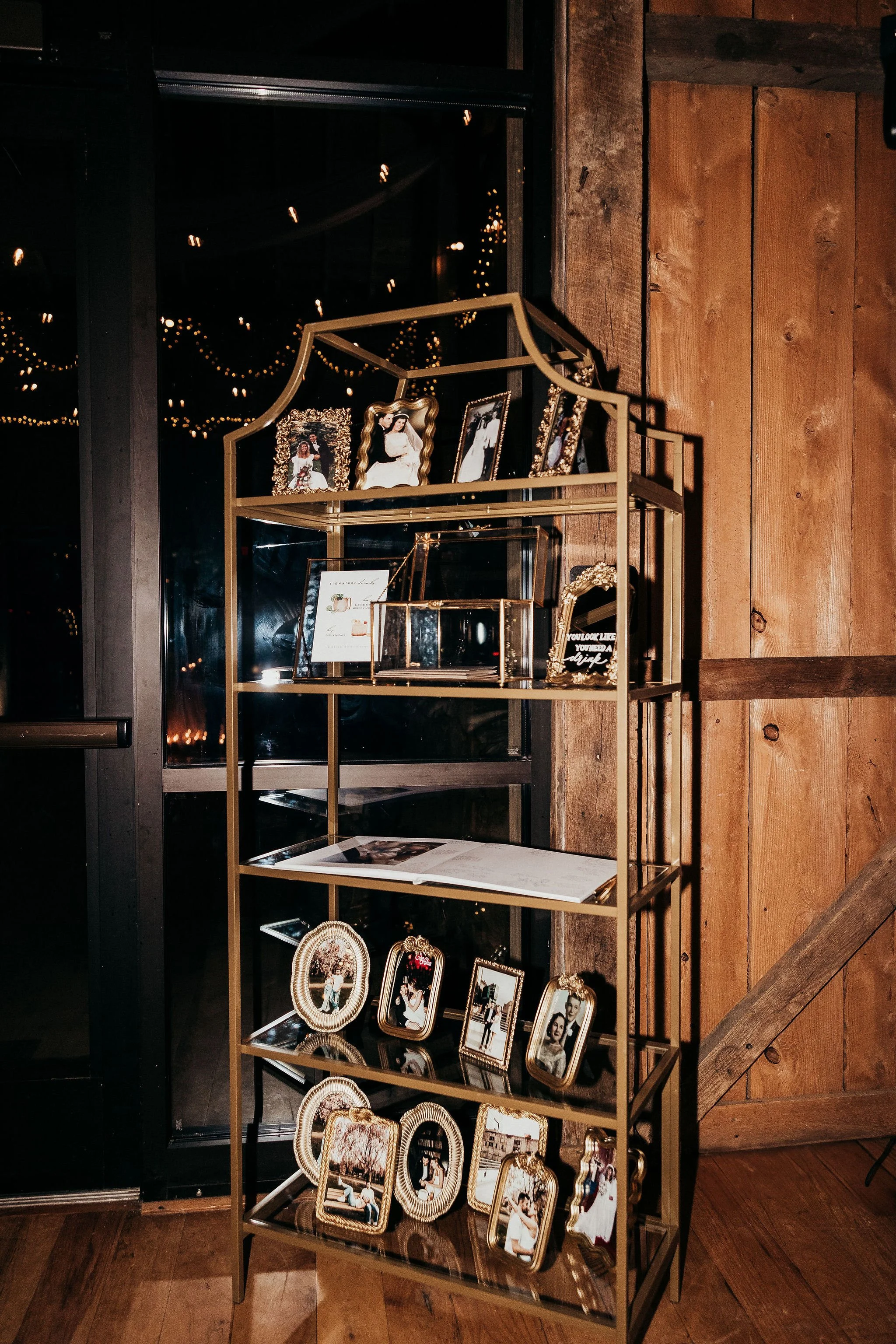 A metal display shelf showing framed photographs and decorative items against a wooden wall with string lights reflected in a window behind it.