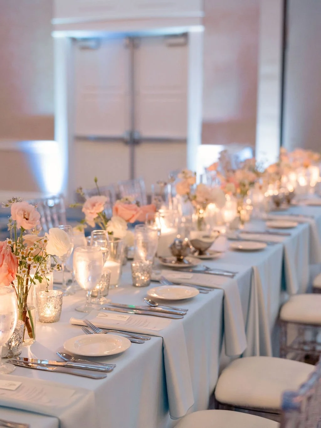 Elegant banquet table decorated with pink and white flowers, candles, and set with plates, glasses, silverware, and napkins, ready for a formal event or wedding reception.