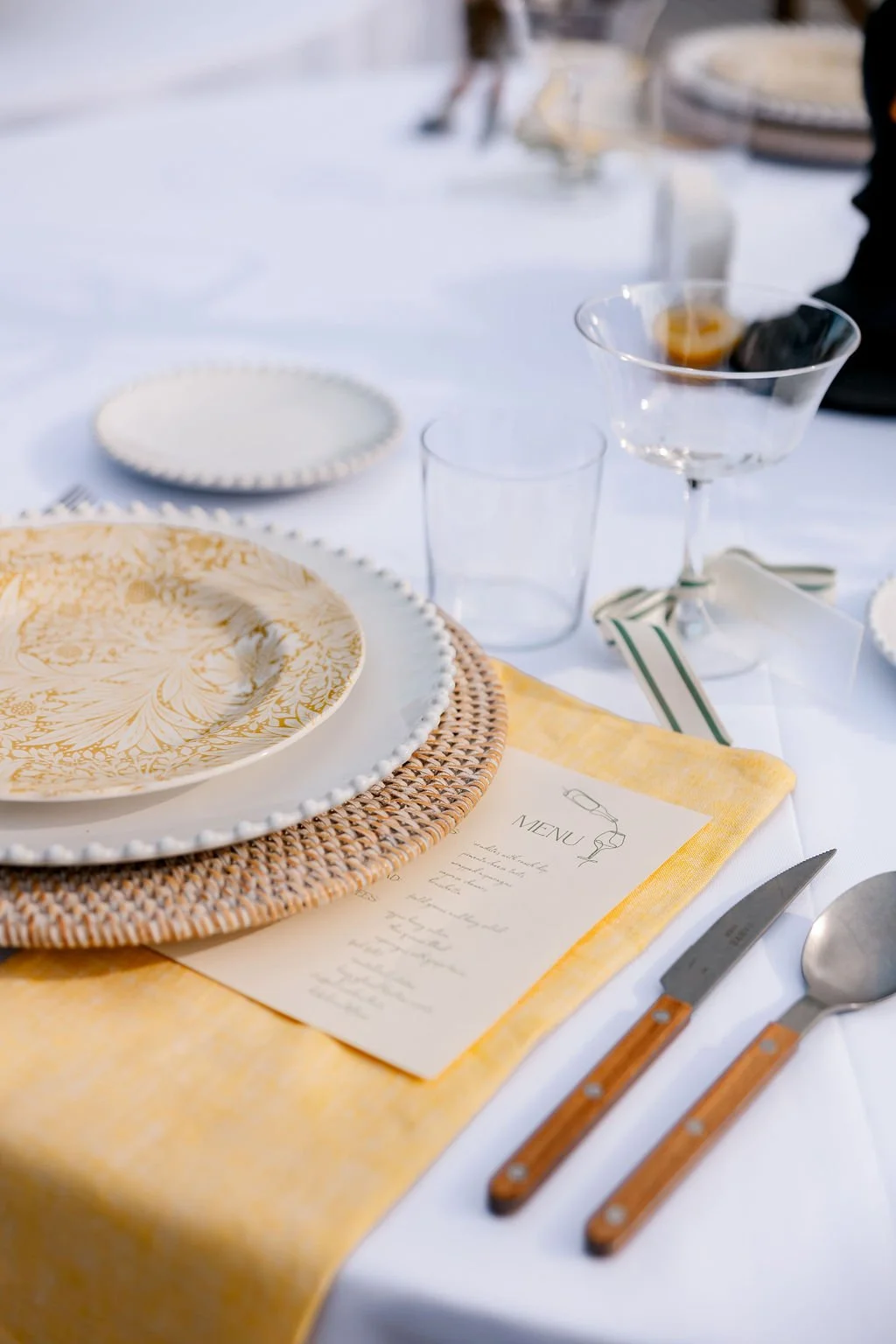 Elegant table setting with a yellow napkin, a white menu card, gold-patterned plate, white and gold-rimmed plate, woven placemat, empty glass, influence cocktail glass with a dark drink, and cutlery with brown handles on a white tablecloth.