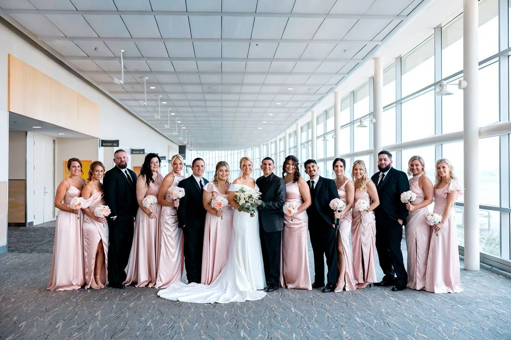 A wedding party of 15 people standing in an airport terminal with large windows, including the bride in a white gown, groom in a black tuxedo, and bridesmaids in pink dresses holding bouquets, posing for a group photo.