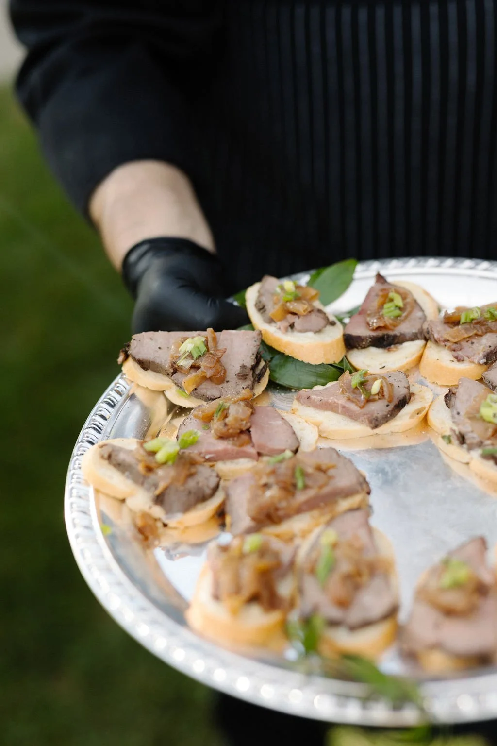 Person wearing black gloves holding a silver tray with assorted open-faced sandwiches topped with meats and garnished with green onions.