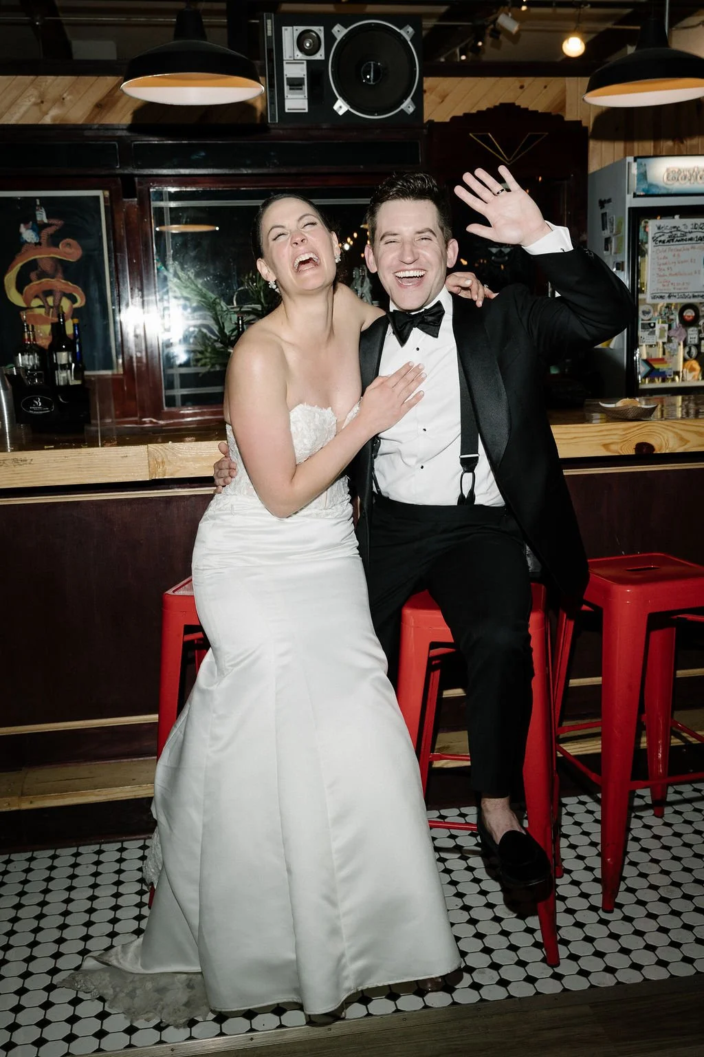 A bride and groom enjoying themselves at a wedding reception, sitting on red bar stools behind a bar, laughing and smiling.