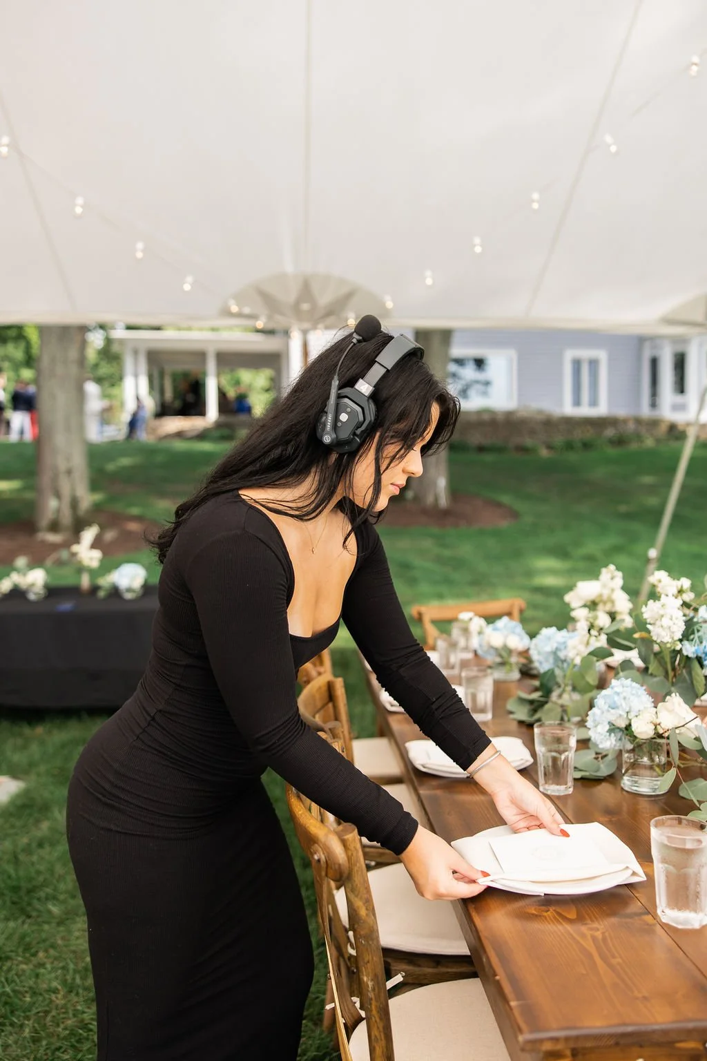 A woman in a black dress setting a table at an outdoor event under a large tent, with floral arrangements and glasses of water on the table.