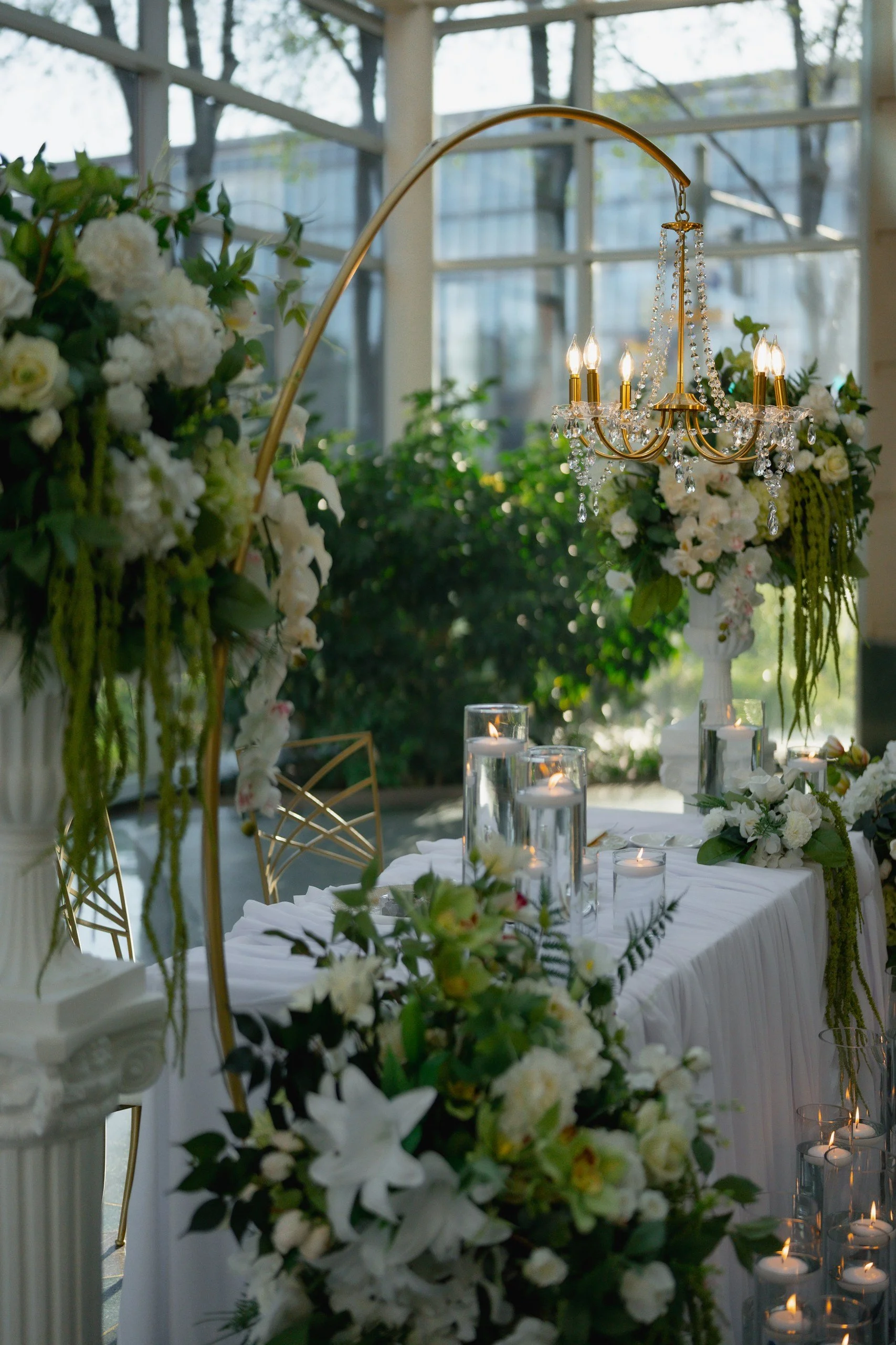A decorated dining table with floral arrangements, candles, and a chandelier inside a bright room with large windows and greenery outside.