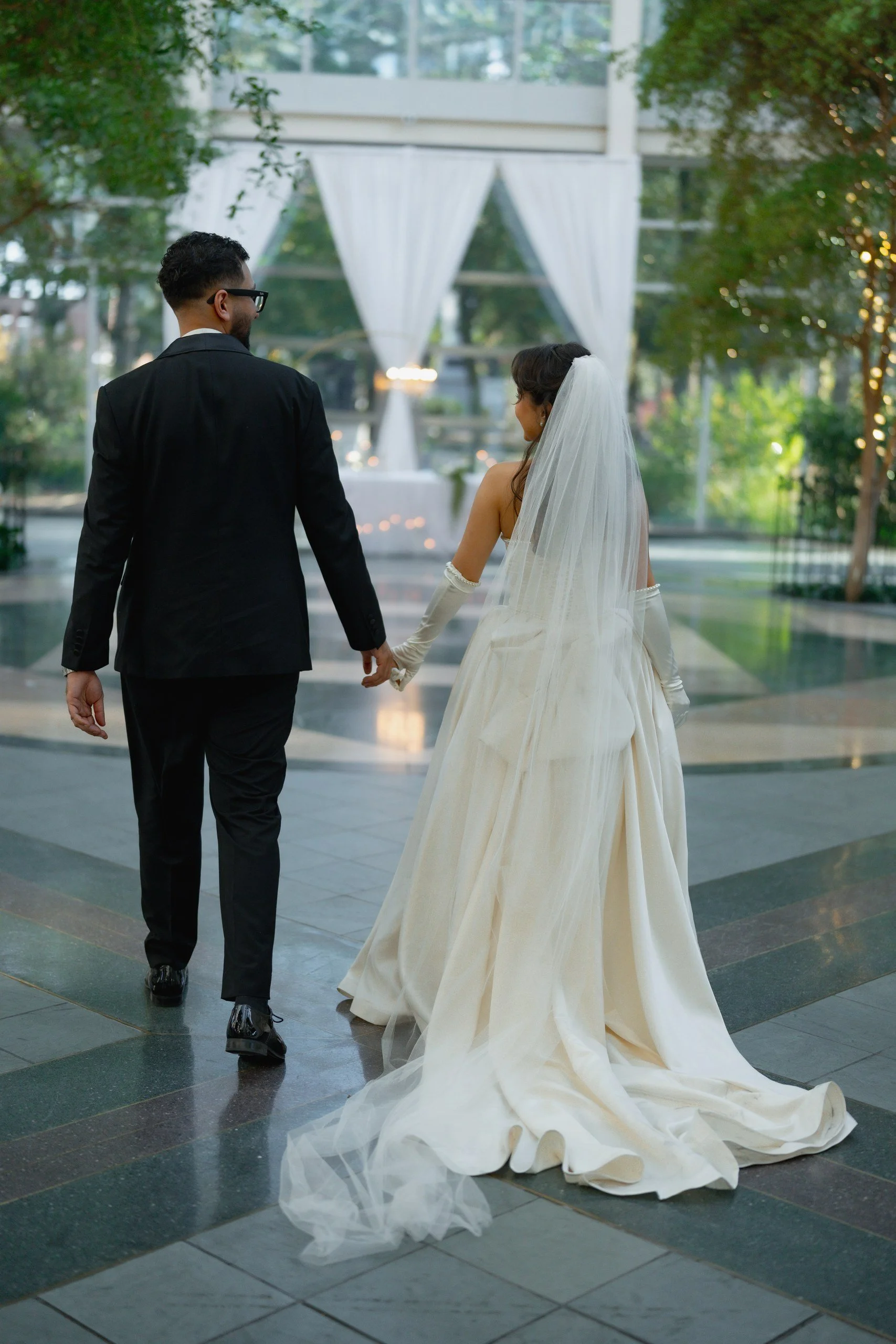 A bride and groom holding hands, walking together indoors with large windows and green trees outside, during their wedding ceremony.
