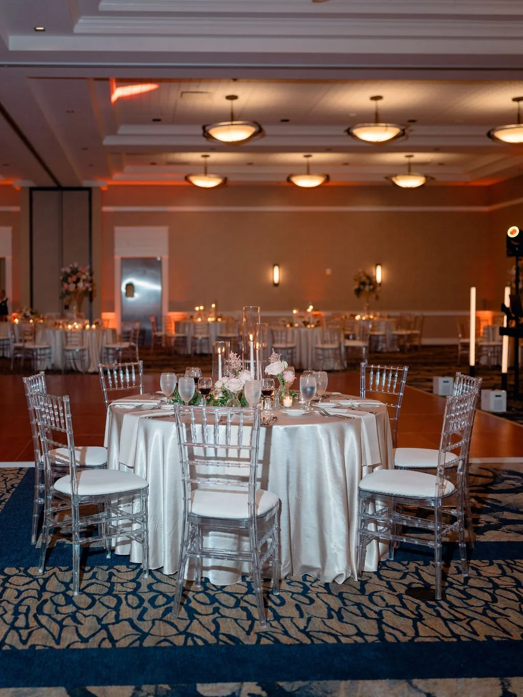 Elegant banquet hall with round table, white satin tablecloth, floral centerpiece, water and wine glasses, and clear acrylic chairs, dim lighting, and decorative wall sconces.