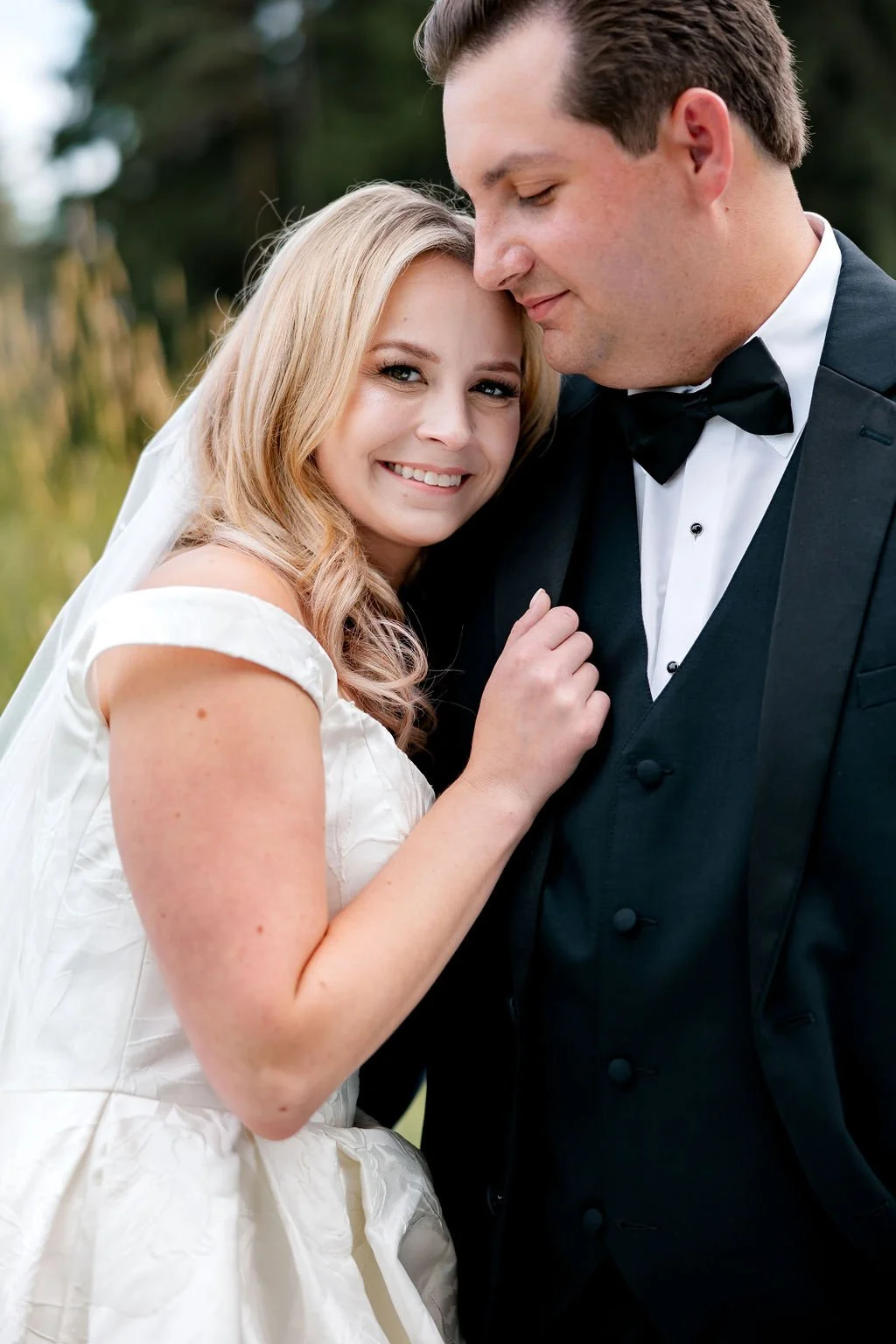 A bride and groom standing close together outdoors, smiling and embracing. The bride is wearing a white wedding dress with off-the-shoulder sleeves and has long, wavy blonde hair. The groom is dressed in a black tuxedo with a bow tie, and has short b