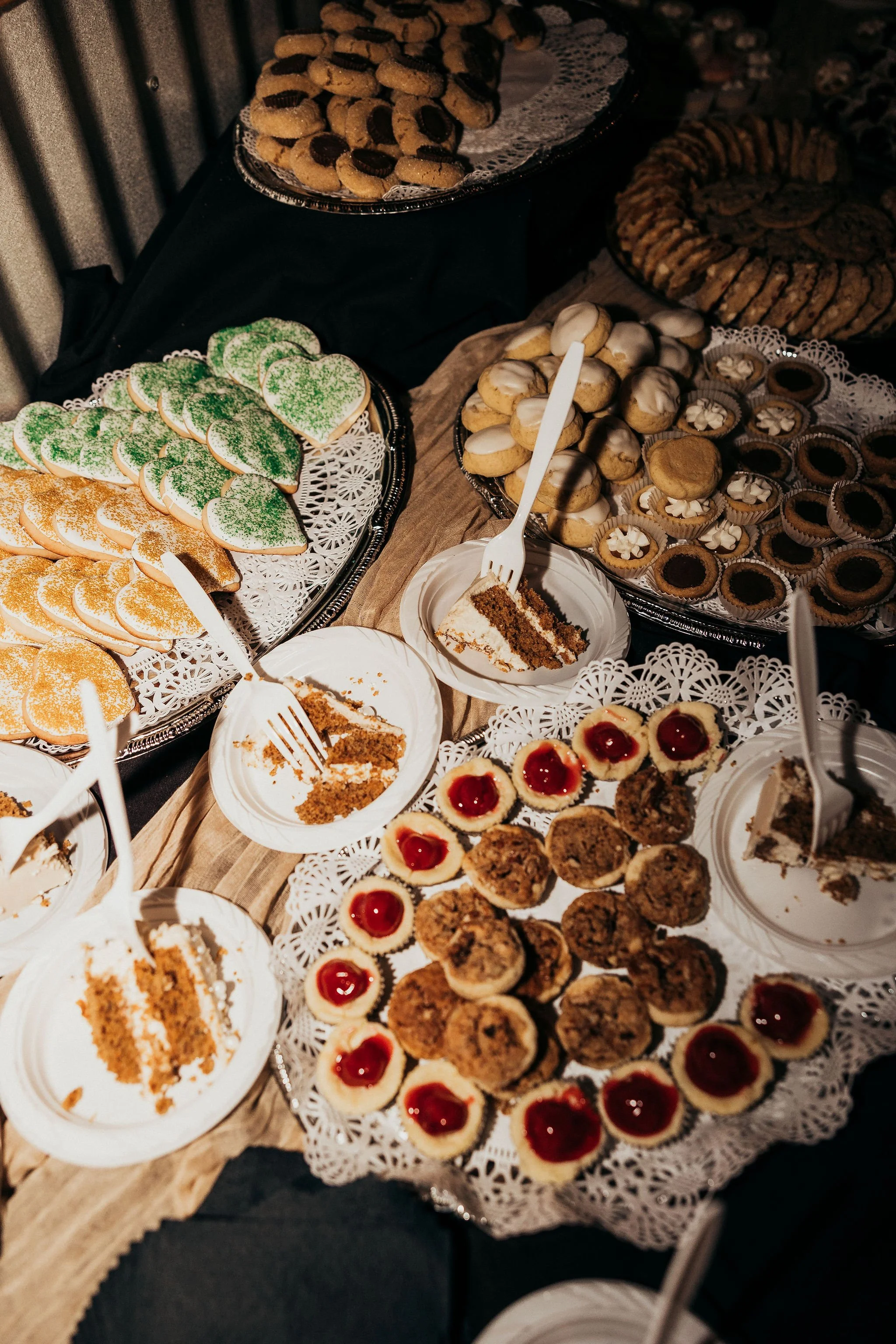 Assorted cookies, including heart-shaped green and orange frosted cookies, chocolate-dipped cookies, and cherry-topped cheesecakes, displayed on trays at a dessert table.