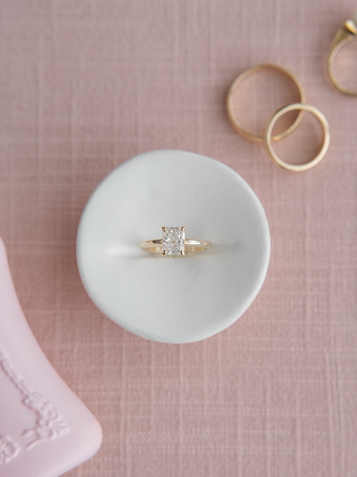 A diamond ring with a rectangular cut stone set in a gold band is displayed on a white circular stand. Several gold rings are arranged in the background on a pink textured surface.