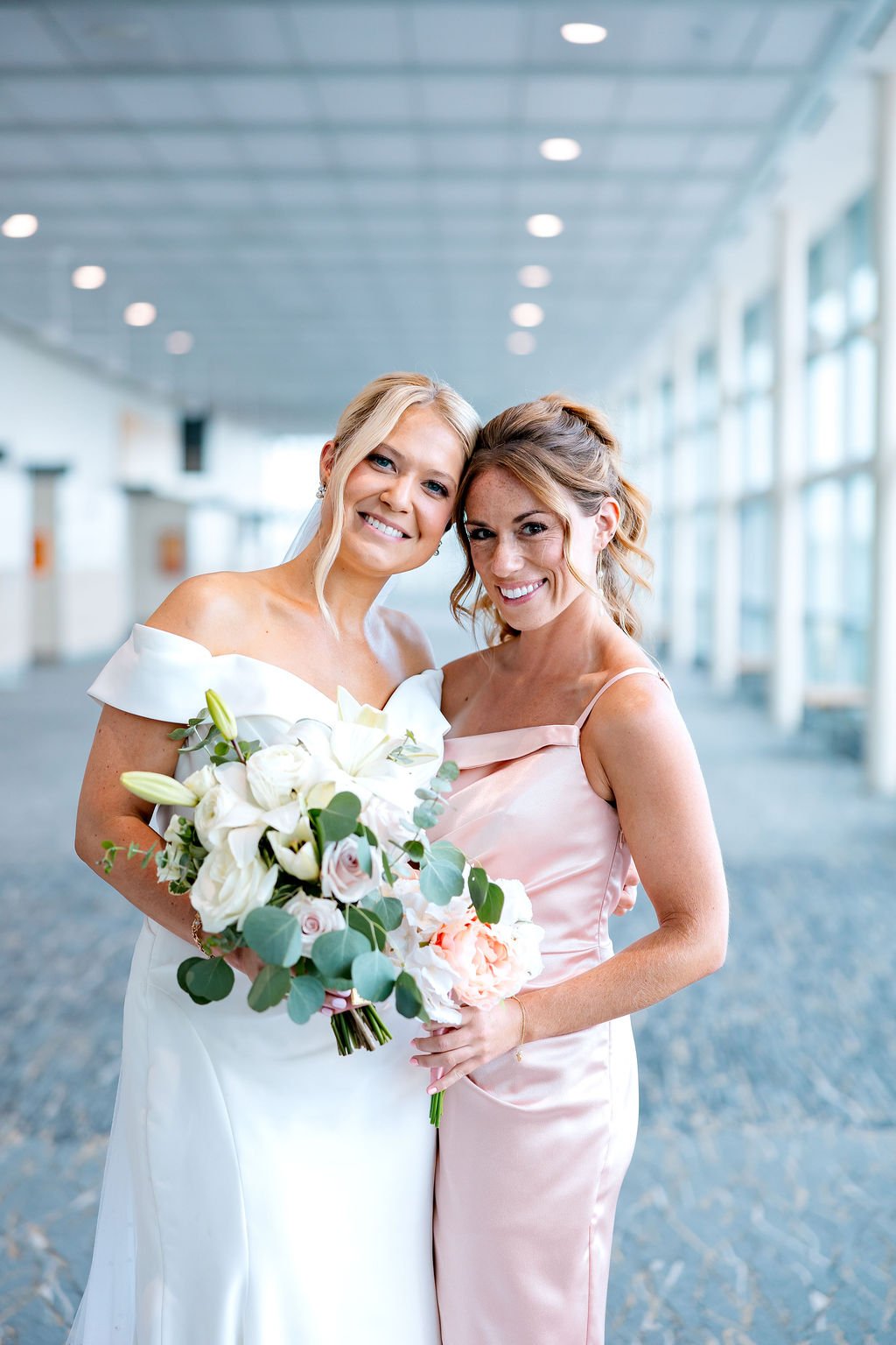 Two women smiling, one in a white wedding dress holding a bouquet of white and blush flowers, the other in a pink satin dress, standing in a bright, modern indoor space.