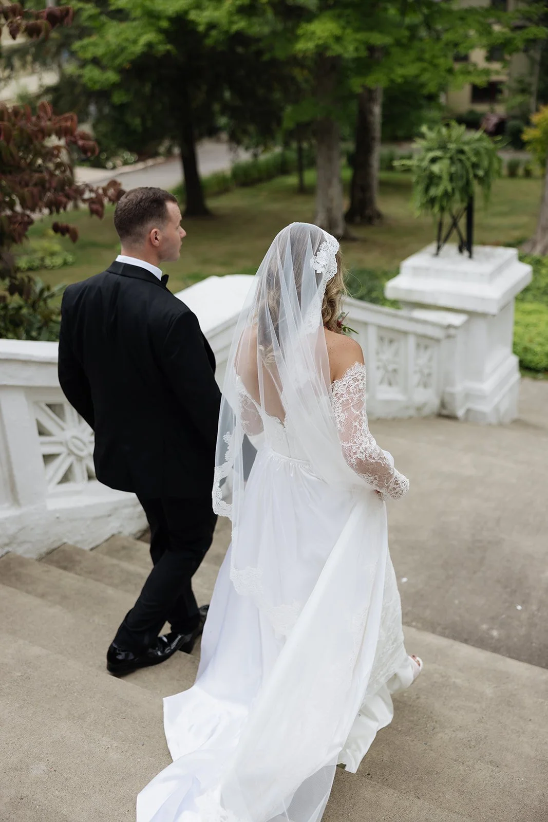A bride and groom standing on stairs outdoors, with the bride in a white wedding dress and veil, and the groom in a black tuxedo, surrounded by trees and greenery.