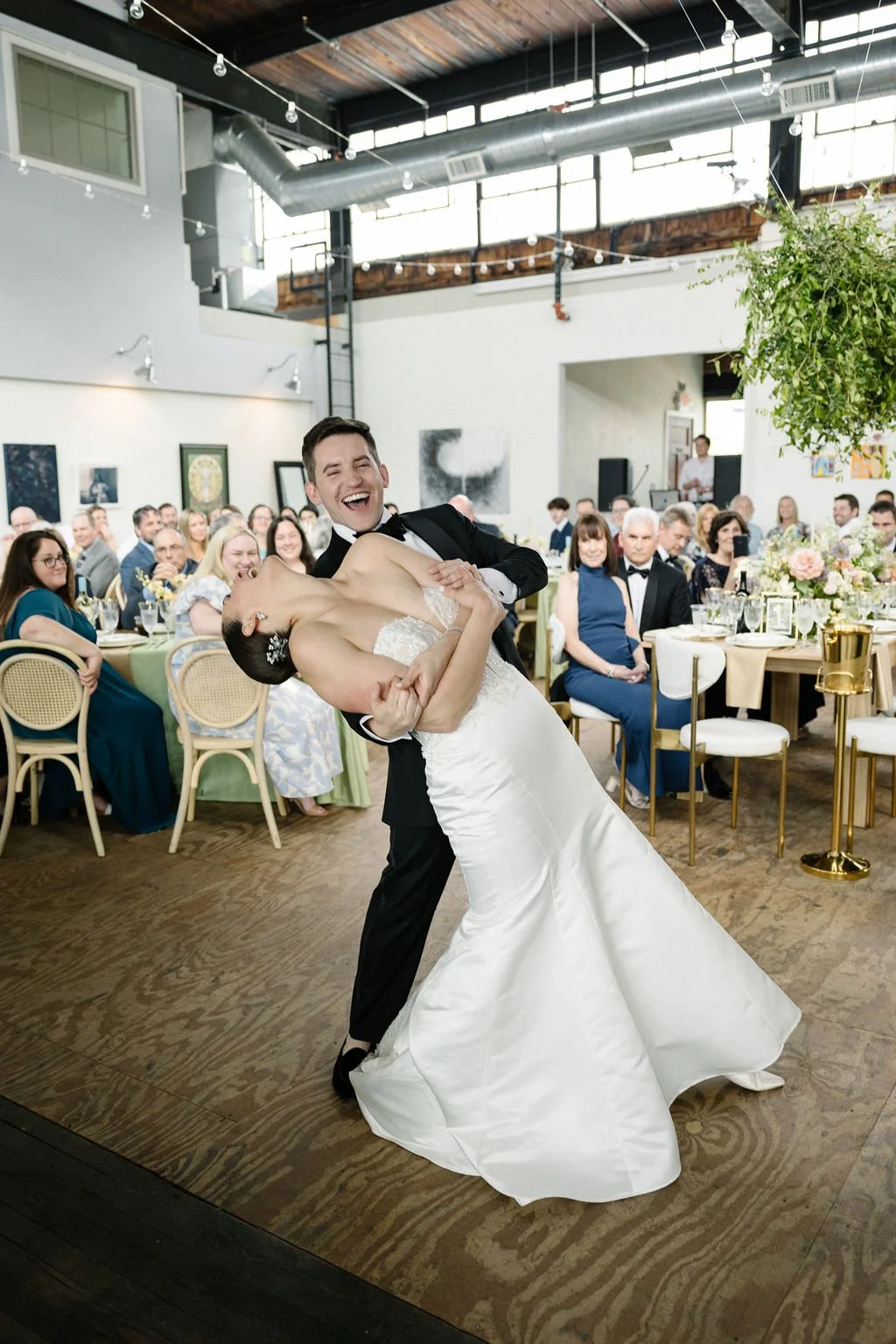 A groom in a tuxedo dancing with a bride in a wedding dress during a wedding reception. Guests are seated at decorated tables in the background, celebrating in a bright, modern venue with high windows and hanging string lights.