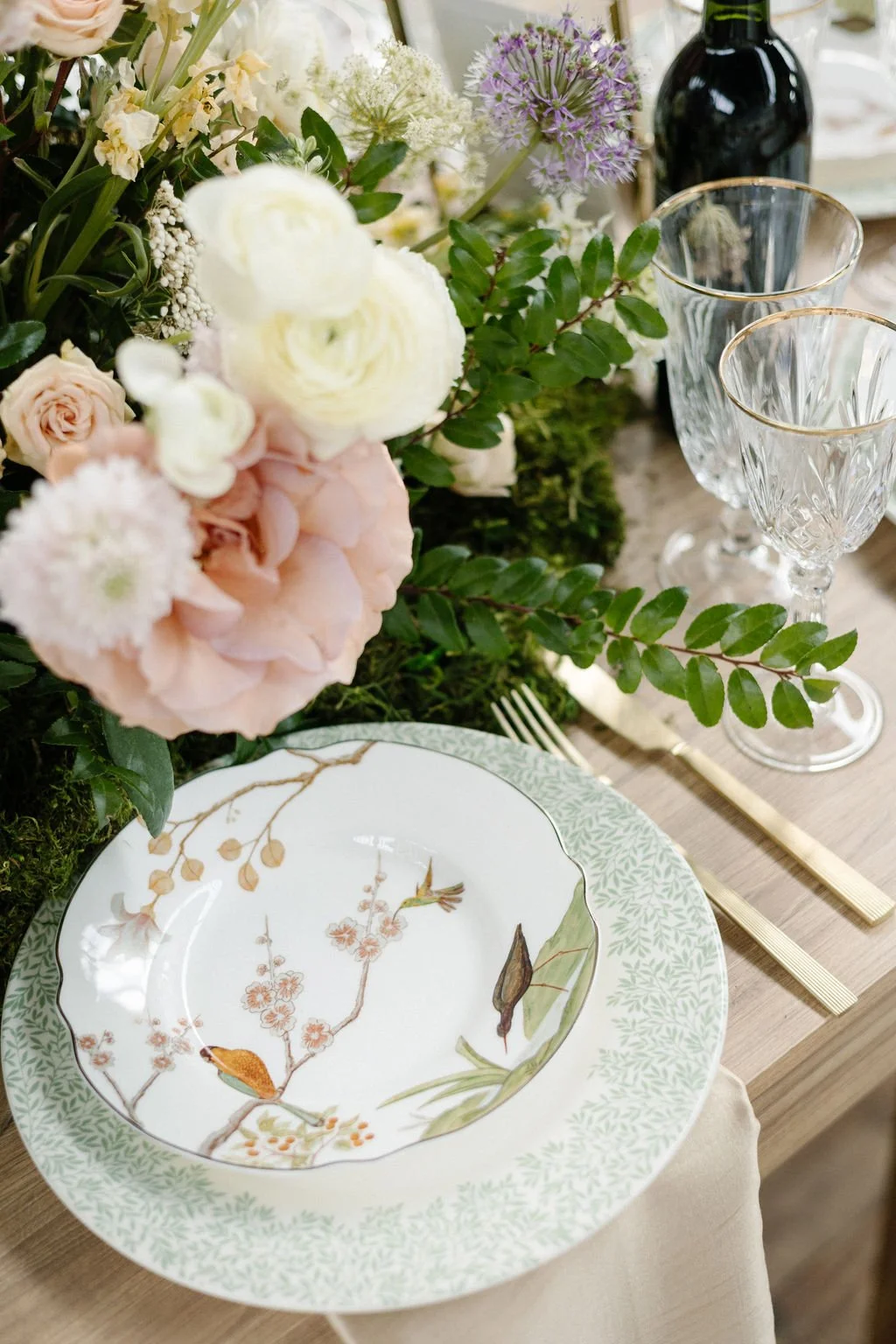 Elegant table setting with floral centerpiece, decorative plates, crystal glasses, gold flatware, and a bottle of wine.