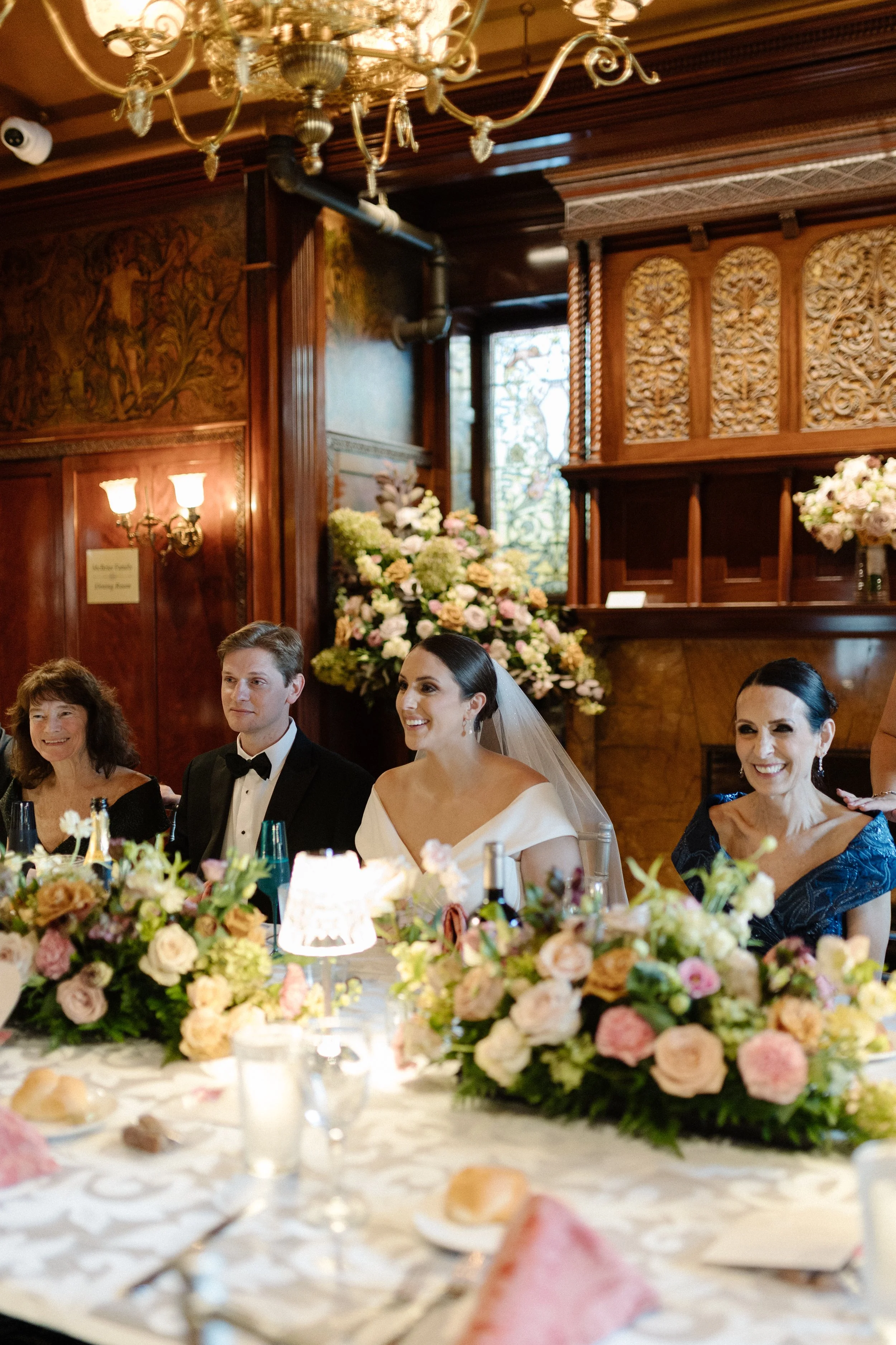 People sitting at a banquet table decorated with pink and white flowers, with a woman in a wedding dress and veil and a man in a tuxedo, in a wood-paneled room with ornate wall decorations and a large floral arrangement in the background.