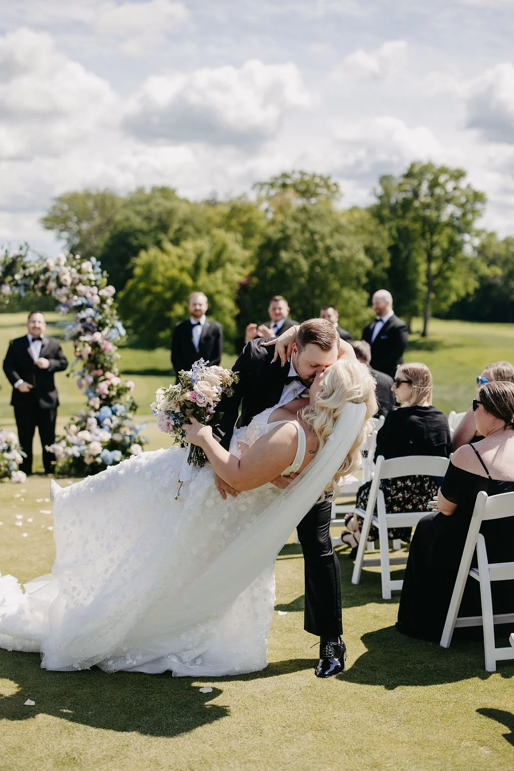 A newlywed couple sharing a kiss during their outdoor wedding ceremony, with the groom dipping the bride while holding a bouquet of flowers, friends and groomsmen in tuxedos in the background on a sunny day.