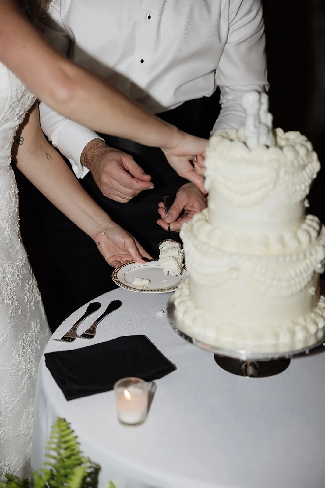 Close-up of a couple cutting a wedding cake at their reception, with a white three-tiered decorated cake on a table, silver forks, a black napkin, and a small candle.