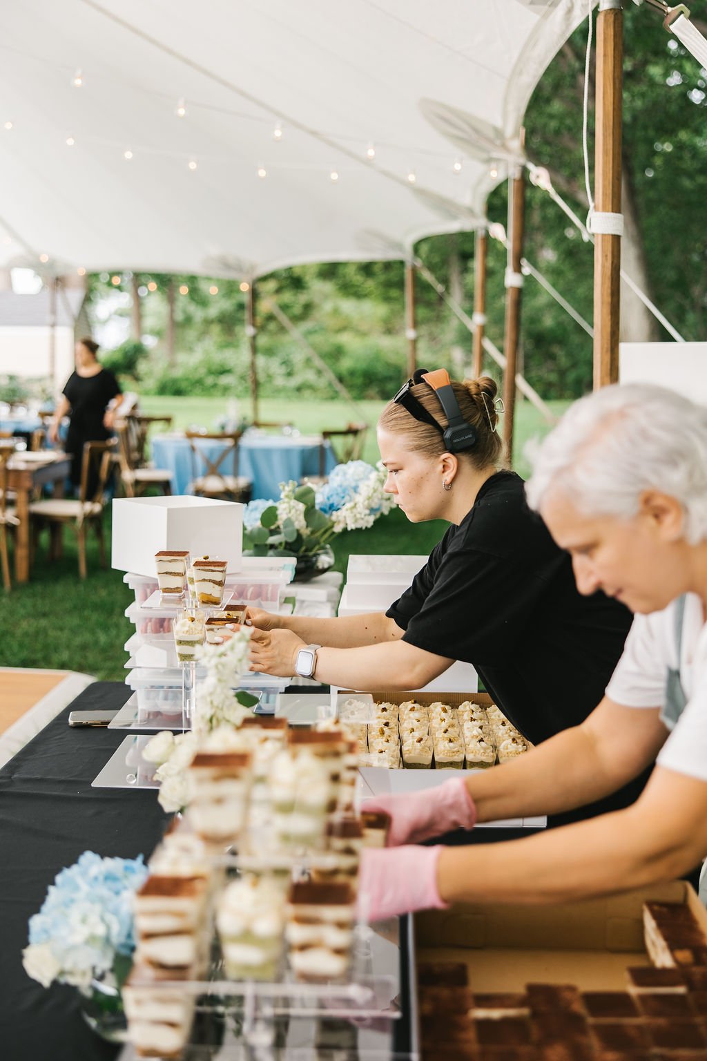 Two women working behind a table filled with assorted layered cakes and desserts under a canopy at an outdoor event.