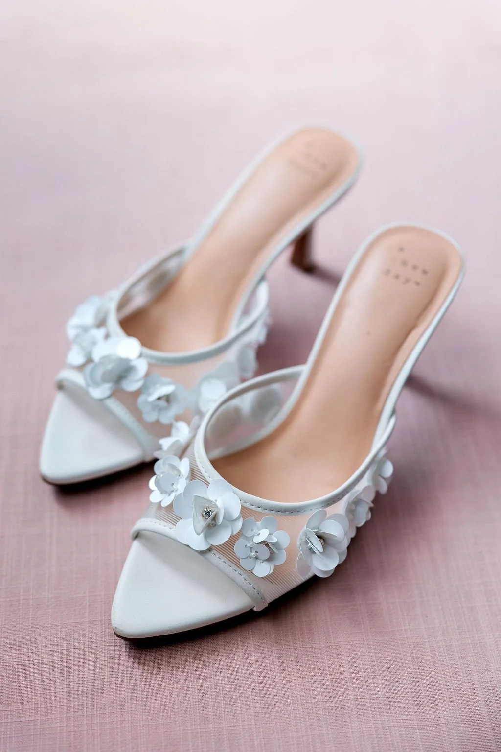 Pair of white high-heeled shoes with floral embellishments on the straps placed on a pink surface.
