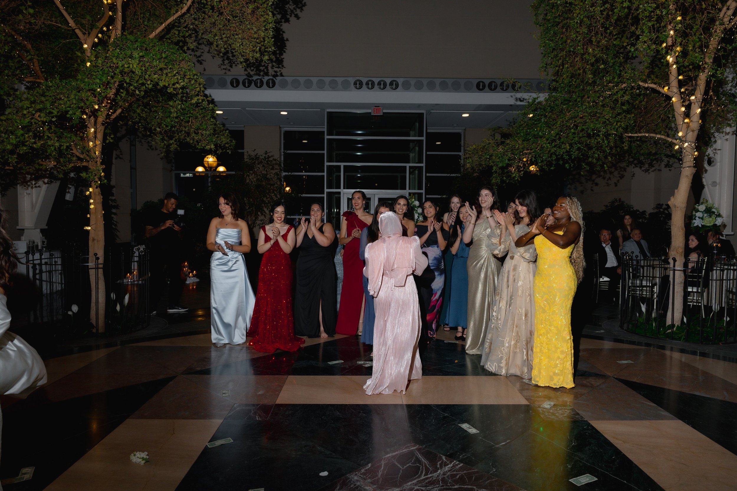 A group of women in elegant dresses standing together at an indoor event, smiling and clapping, with some people seated in the background.