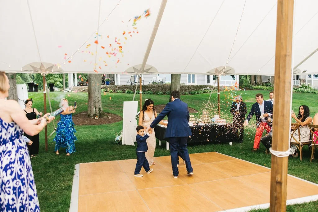 People celebrating at an outdoor wedding reception with confetti, under a large white tent on a grassy lawn.
