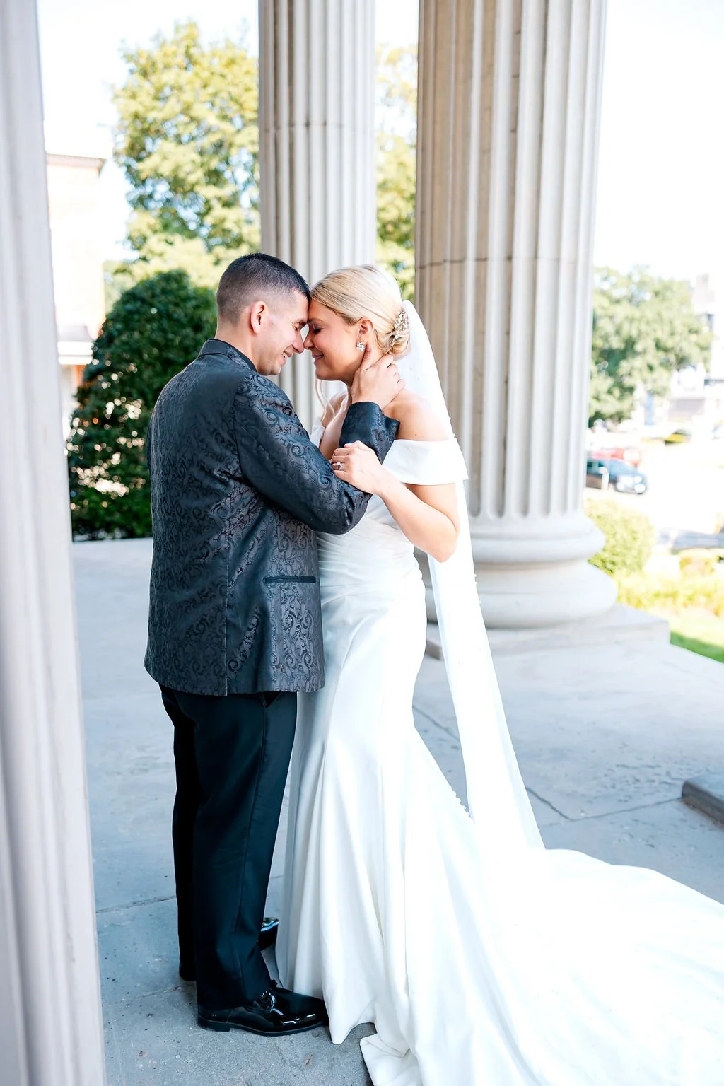 A bride and groom share a tender moment on their wedding day, touching foreheads and smiling, surrounded by classical columns and greenery.