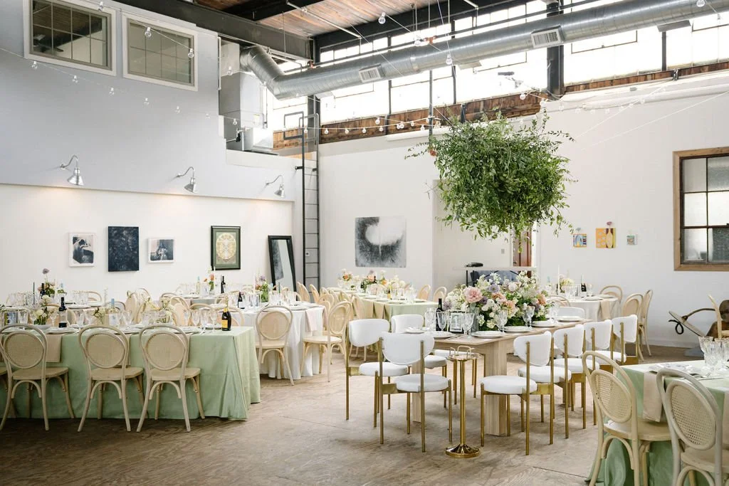 A decorated event space with round tables covered in pastel-colored tablecloths, surrounded by white and beige chairs. The tables have floral centerpieces and place settings, and the room features hanging string lights, artwork on the walls, and a la
