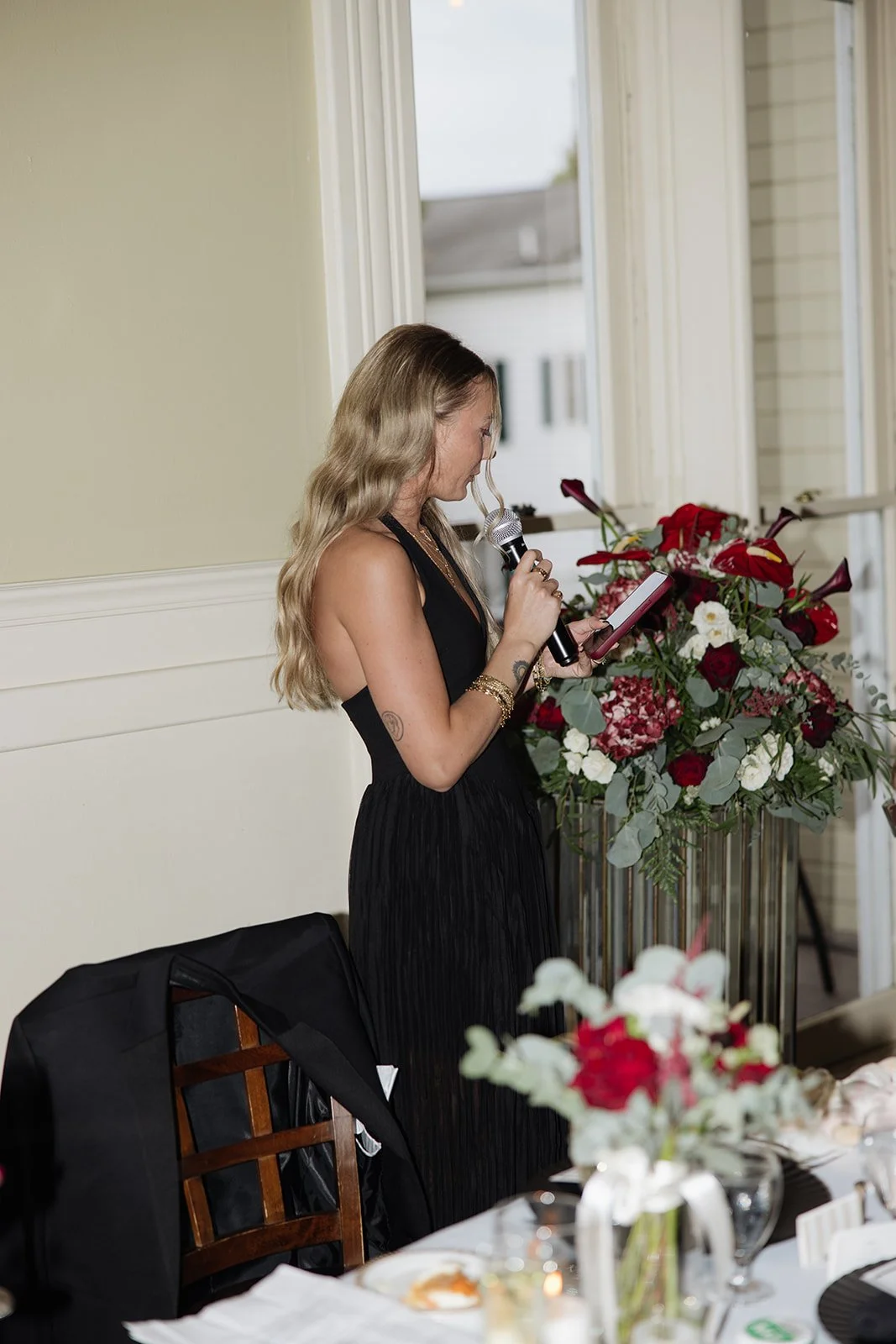 A woman with long, wavy blonde hair, wearing a black sleeveless dress, stands at a table holding a microphone in one hand and a tablet device in the other, speaking or reading. The table area is decorated with a large floral arrangement of red and wh
