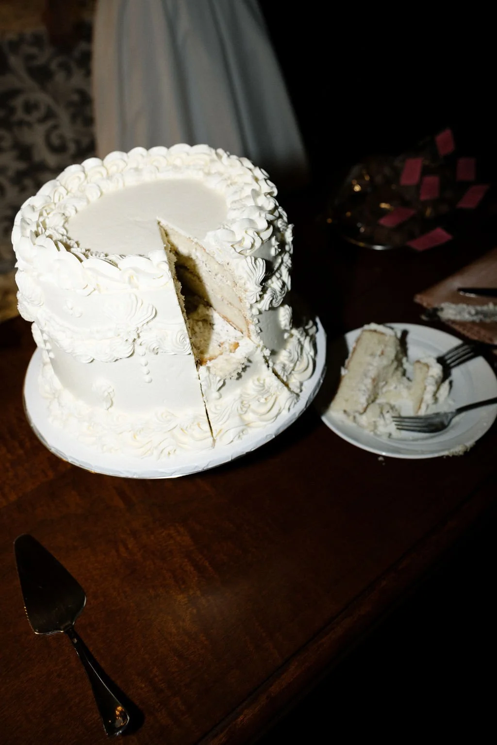A white wedding cake with decorative piping and a slice missing, placed on a wooden table. A plate with a slice of cake and a fork are next to it.